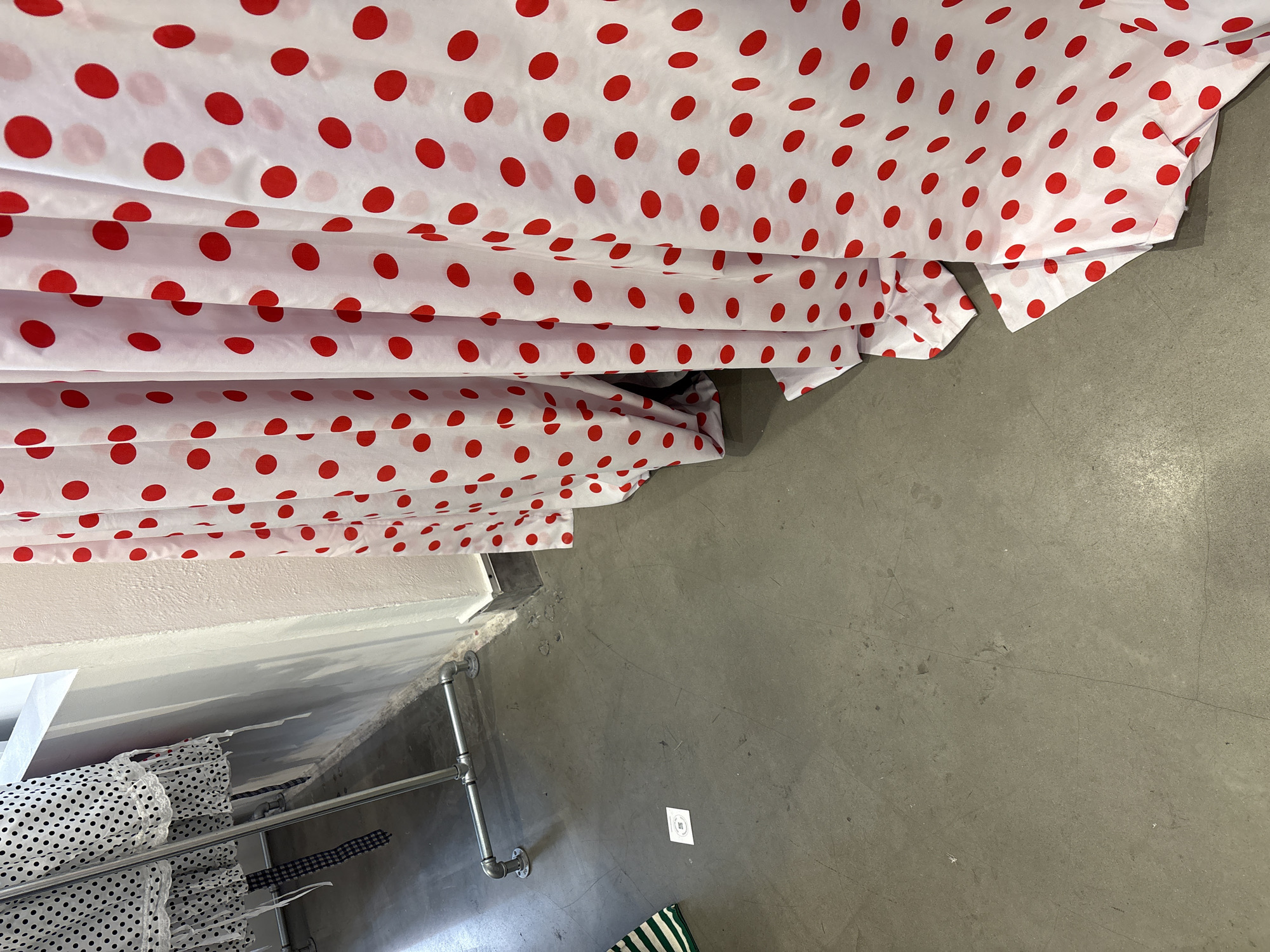 The image depicts a clothing store interior, likely in London, United Kingdom, based on common architectural elements. The view is from an elevated angle, looking down towards a changing room area. Dominating the upper left is a stack of folded white fabric with red polka dots, possibly curtains or clothing items. Adjacent to the polka dot fabric is a gray, concrete floor extending into the right portion of the image. On the left side, a clothing rack made of metal pipes is visible, holding garments with black and white polka dot patterns and checkered patterns. A strip of white wall runs along the top of the clothing rack. On the floor, near the bottom of the image, there is a green and white striped object, perhaps a bag or mat. A small white sign with faint markings lies on the concrete floor. The overall impression is of a well-lit retail space with organized clothing displays.