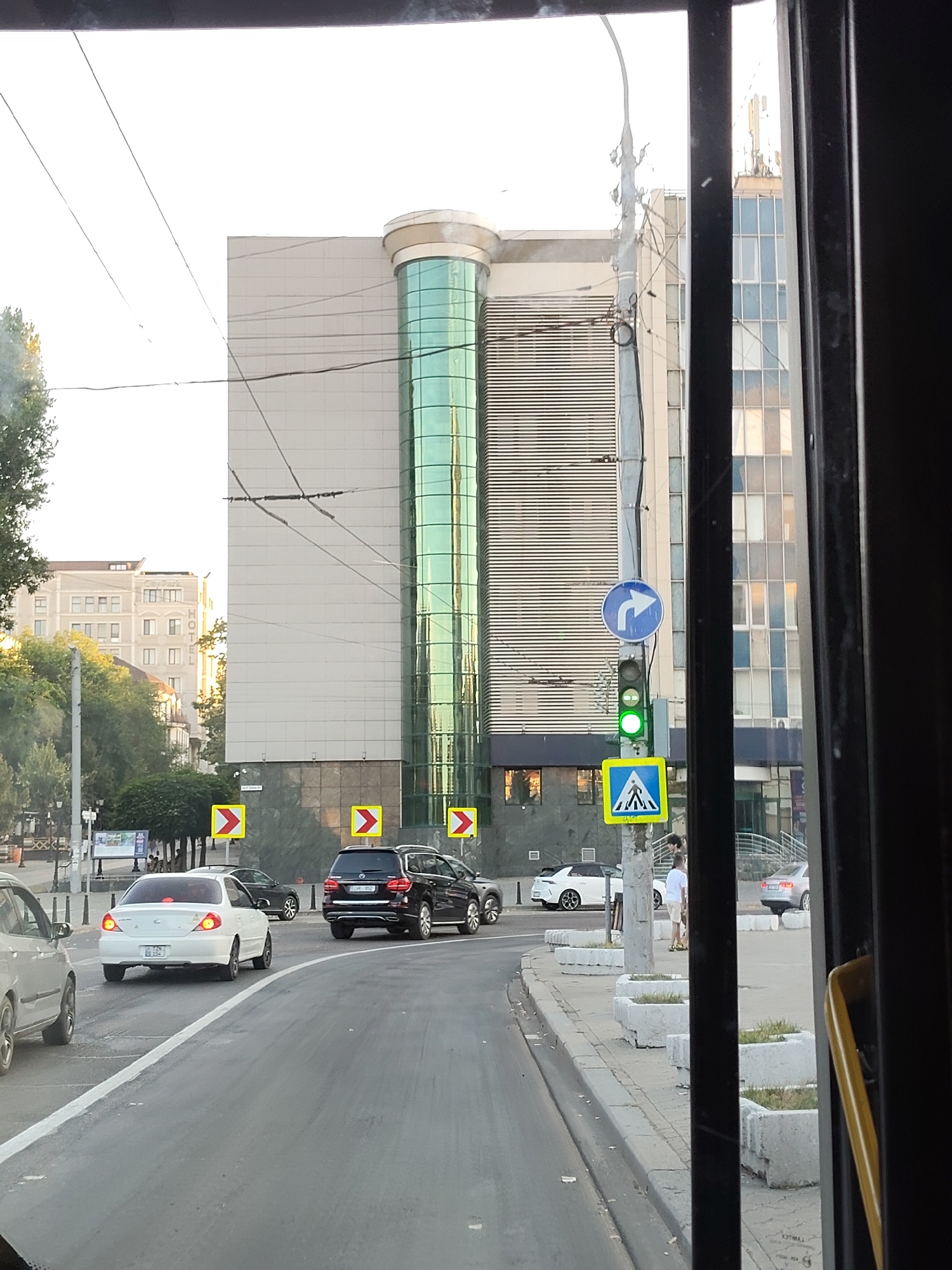 The image shows a street scene in Chișinău, Moldova, likely captured from inside a vehicle. In the foreground, the road curves to the right, with several cars present. A white sedan is in the center, its brake lights illuminated. To its left, a portion of a light-colored car is visible. Further ahead and to the right, a black SUV is also on the road.
The background features a modern, multi-story building with a distinctive glass-covered cylindrical section. Adjacent to this building, another taller building with a grid-like facade is visible. A traffic light, showing green, is mounted on a pole along with traffic signs. One sign indicates a right turn is permitted, and another is a pedestrian crossing symbol. Several yellow directional signs with red arrows pointing to the right are also present along the roadside. The environment appears to be a business or commercial district, with trees and sidewalk planters lining the street. The sky is bright, suggesting daytime.