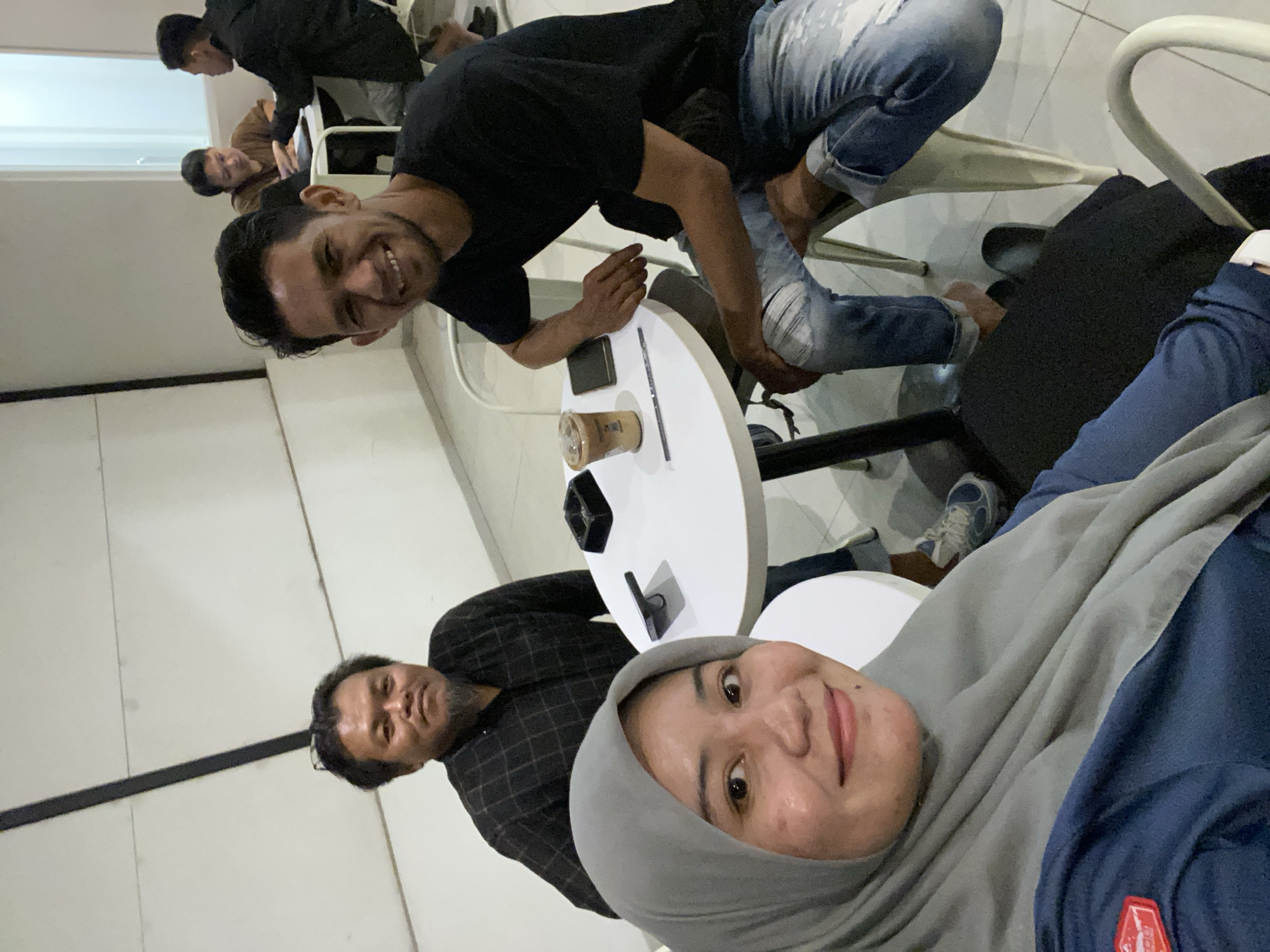 The image captures a candid moment in what appears to be a cafe or casual dining establishment in Banjarbaru, Indonesia. In the foreground, a woman wearing a grey hijab and a dark blue shirt with a red and white badge on the left sleeve smiles at the camera. Behind her, a man with dark, medium-length hair and a goatee, wearing a black checkered shirt, is also looking towards the camera. To the right, another man with short, dark hair and a beard, dressed in a black t-shirt, beams at the viewer. He is seated at a white circular table, leaning forward with his left arm resting on his lap. On the table, a cup with a straw and a phone are visible, along with an ashtray. In the background, two other individuals are seated at another table, partially obscured. The environment features white walls and simple white chairs, suggesting a modern and uncluttered setting. The lighting is soft, creating a warm and relaxed ambiance.