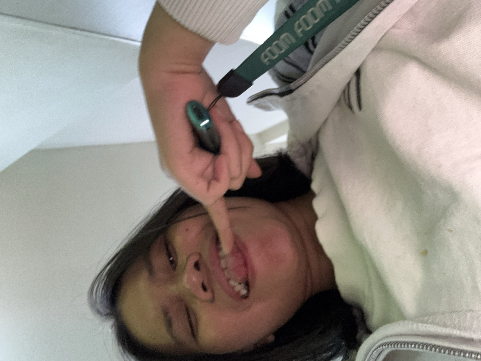 In a dimly lit interior setting, possibly a room with white walls, a young East Asian woman is captured from a low, upward angle. She has dark hair that falls around her face and shoulders. Her eyes are partially closed, and she is pulling on her lower lip with her right index finger, revealing her teeth. She is wearing a light-colored, possibly white, crewneck sweatshirt and an open light gray zipper jacket over it. A green lanyard with the repeated word "FOOM" printed in white is attached to the jacket. She is holding a small, dark green electronic device with a metallic trim in her right hand, near her mouth. The overall lighting suggests an indoor environment, potentially a residential or personal space, and given the context of East Jakarta, Indonesia, it is likely this is a private dwelling.