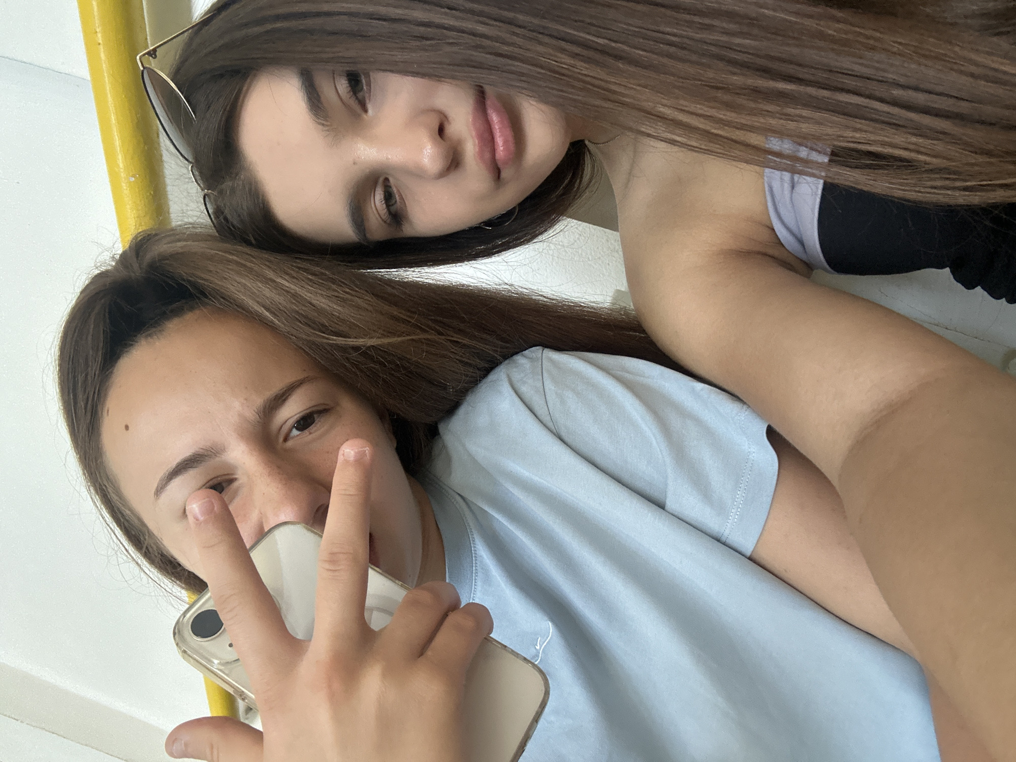 Two young women are captured in a selfie, their faces close to the camera. The woman in the foreground, closer to the viewer, is holding a smartphone in her right hand, obscuring part of her face. Her left hand is raised, with two fingers extended in a peace sign. She has long, brown hair and is wearing a light blue t-shirt. The woman behind her, slightly out of focus, has her head resting on the other woman's shoulder or arm. She has long, dark brown hair and is wearing sunglasses. Her expression appears relaxed, with her eyes looking slightly downwards. The background is a plain, light-colored wall, and a bright yellow pole is visible to the left. The lighting suggests it is daytime, likely indoors or in a shaded outdoor area. The overall mood seems casual and friendly, with the two women appearing to be enjoying each other's company. The image does not contain any visible text.