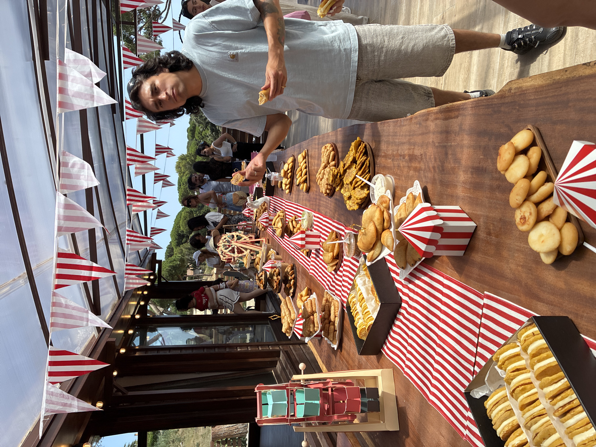 The image depicts an outdoor buffet table laden with various food items, set against a backdrop of a wooden building with large windows and a clear, sunny sky. Red and white striped pennant banners are strung overhead, creating a festive atmosphere. The table itself is covered with a red and white striped runner.
On the table, several platters and containers hold an assortment of pastries, fried foods, and what appear to be empanadas. To the left, a small, colorful Ferris wheel model and a taller, multi-tiered model of a carnival ride are visible. In the foreground, a man with dark, wavy hair is holding a piece of food in each hand, seemingly about to eat. He is wearing a light blue t-shirt and grey shorts. Other people are visible in the background, mingling and looking at the food. The setting appears to be a casual outdoor event. The presence of a Ferris wheel model and carnival-themed decorations suggests a fair or celebration. The location is identified as Sant Vicenç De Montalt, Spain, which implies a Mediterranean setting, possibly a garden or patio area associated with a venue.