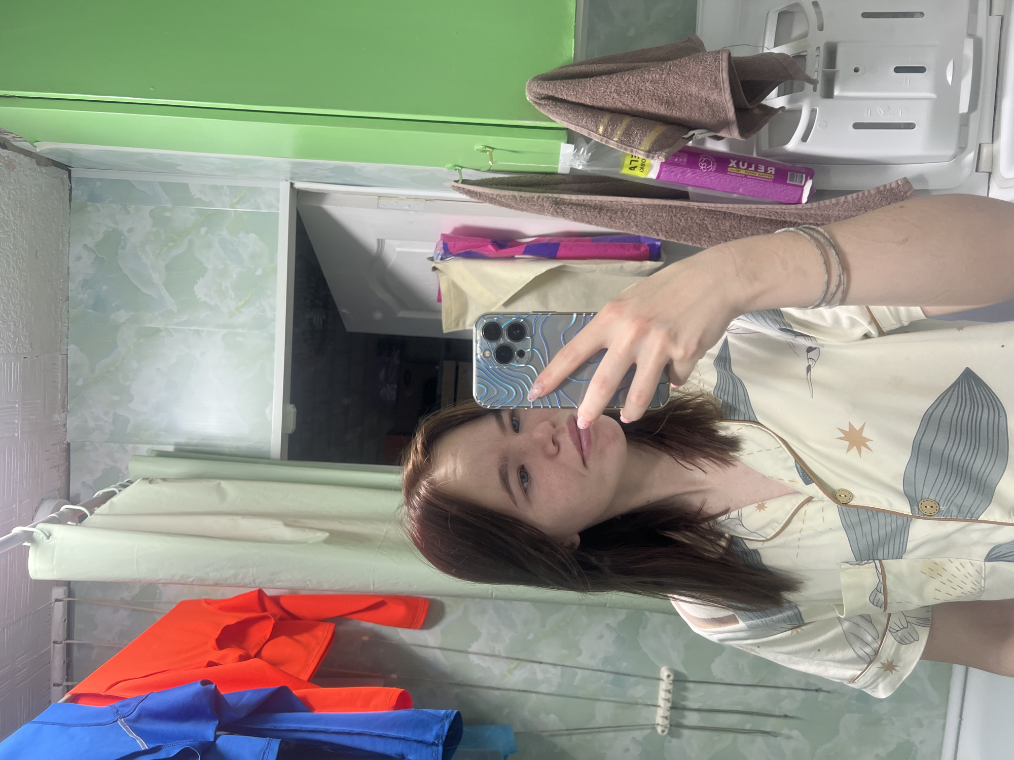 A person is taking a selfie in a bathroom. The person has brown hair and is wearing a patterned pajama shirt. The bathroom has light green and white marble-patterned tiles on the walls. There is a drying rack with clothes hanging on it, including an orange shirt and blue garments. Above the person, there is a green cabinet. A brown towel and some toiletries are placed on top of the cabinet. The location is a bathroom, likely in the city of Chaikovsky, Russia.
