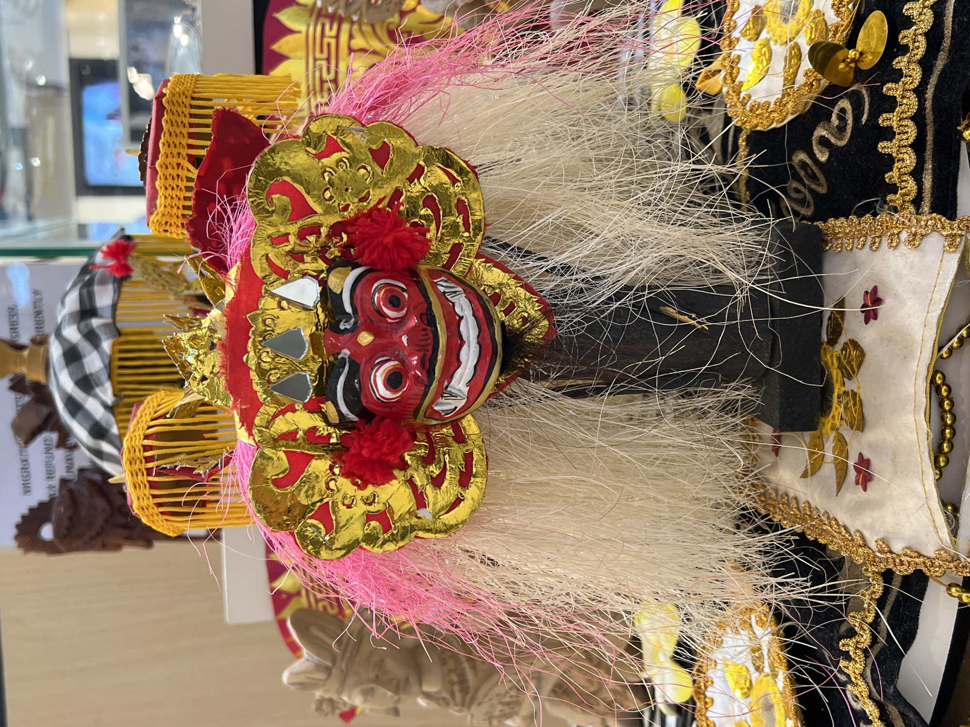 This image shows a traditional Balinese Barong mask, likely a tourist souvenir, given its pristine condition and display setting. The mask features a red face with bulging white eyes and a prominent white fanged smile, framed by black details. Elaborate gold and red adornments surround the face, accented with small mirrors. Attached to the mask is a thick, flowing mane of white and pink fibers simulating hair. The mask is set against a backdrop that includes other Balinese artifacts and tourist items, suggesting the location is a souvenir shop, perhaps within the city of Denpasar, Indonesia, given the prevalent Balinese cultural items. The arrangement is likely part of a retail display aimed at tourists.