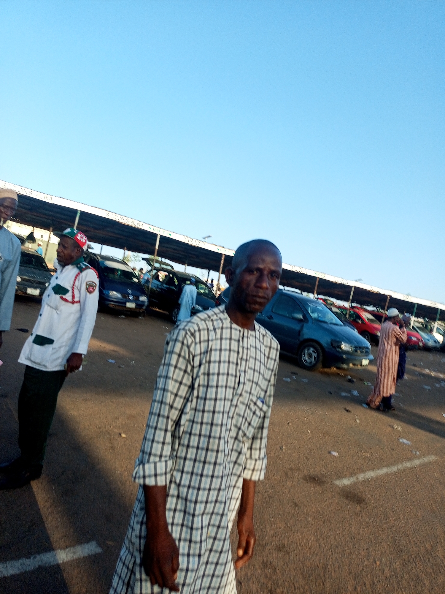 The image depicts several individuals in an outdoor setting, seemingly a parking lot or open area under a covered structure. The sky is clear and blue.
The primary subject is a man in the foreground wearing a plaid robe. To the left, another man is partially visible, dressed in a white uniform-like attire with green and red details. Other people are scattered in the background, some wearing traditional clothing. Cars of various colors and models are parked in rows behind the individuals.
The ground is paved, and there appears to be some debris or litter present.
