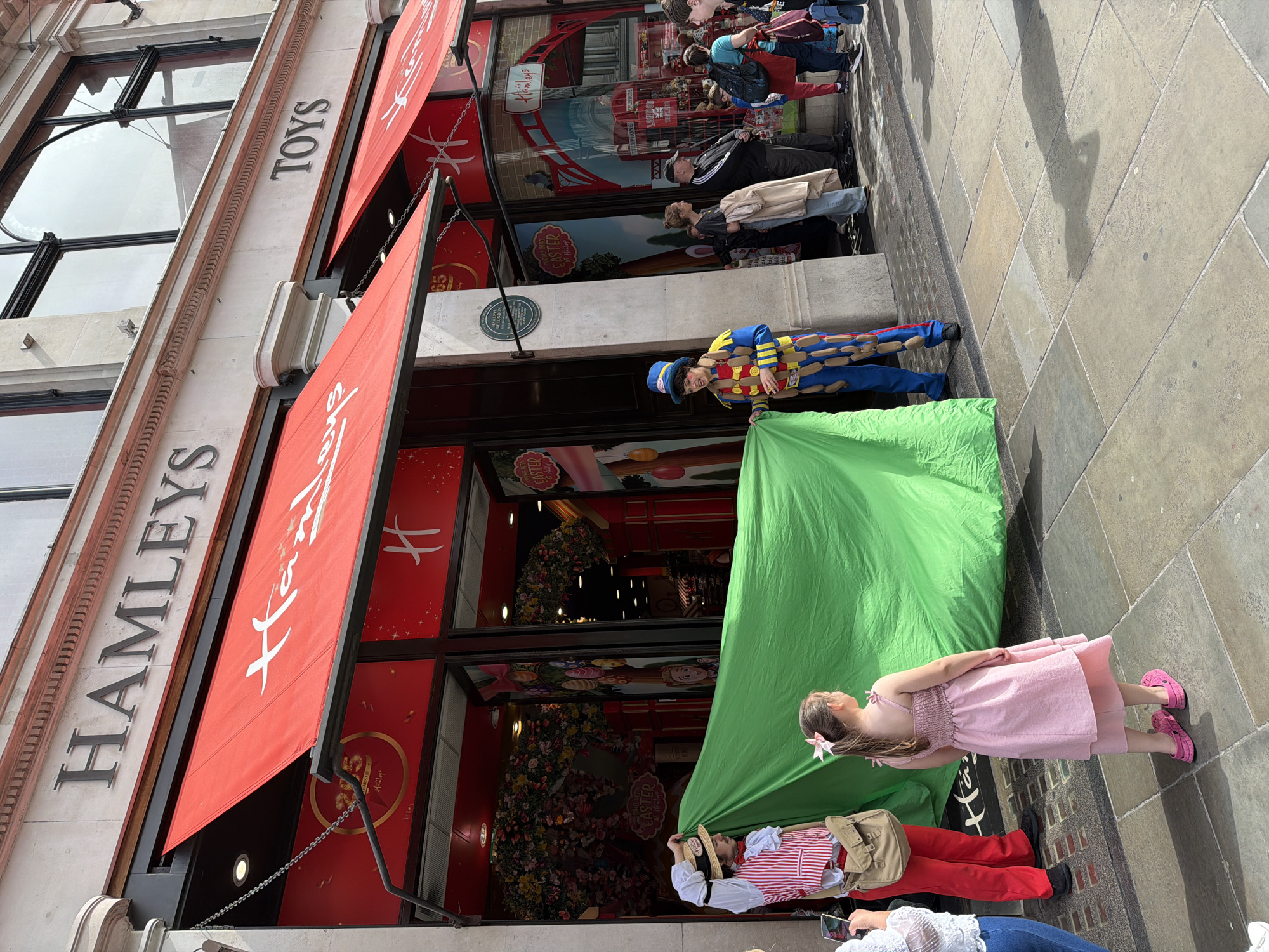The photograph depicts the exterior of Hamleys toy store in London, United Kingdom. The store has a red awning and large windows displaying toys and Easter decorations. A clown and a person in a traditional outfit are holding up a large green sheet. A young girl in a pink dress is standing next to them. Several people are walking by on the sidewalk.
