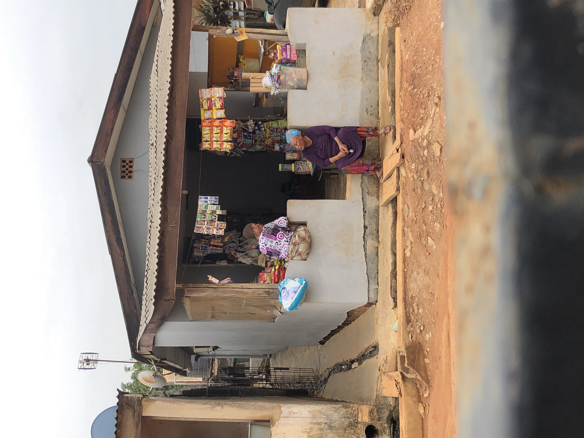 This outdoor image captures a vibrant, open-front market stall or small shop, likely in a bustling community. The shop is housed in a simple building with whitewashed walls and a corrugated metal roof, showing signs of wear and age, including cracks in the concrete steps and rough-textured walls. Various packaged goods, snacks, and possibly toys are displayed on shelves and hanging from the storefront, indicating a local convenience or grocery store.
Two women are present. One woman, dressed in a purple outfit and a blue head covering, is seated on a concrete ledge outside the shop, holding a small object in her hands and seemingly looking down at it. She is positioned towards the right side of the shop entrance. Further inside the shop, a second woman is visible, wearing a colorful, patterned top with the word "ROCK" prominently displayed on it. She appears to be attending to the goods or resting. Near her is a light blue bag featuring a printed face.
The immediate foreground consists of a reddish-brown, unpaved ground, suggesting dirt or rough earth. In the background, under a bright, overcast sky, architectural elements like a satellite dish and a tall, slender antenna with a cage-like structure are visible, indicating the presence of communication infrastructure. The scene suggests a typical daytime operation of a small, local business, serving the community with everyday items.