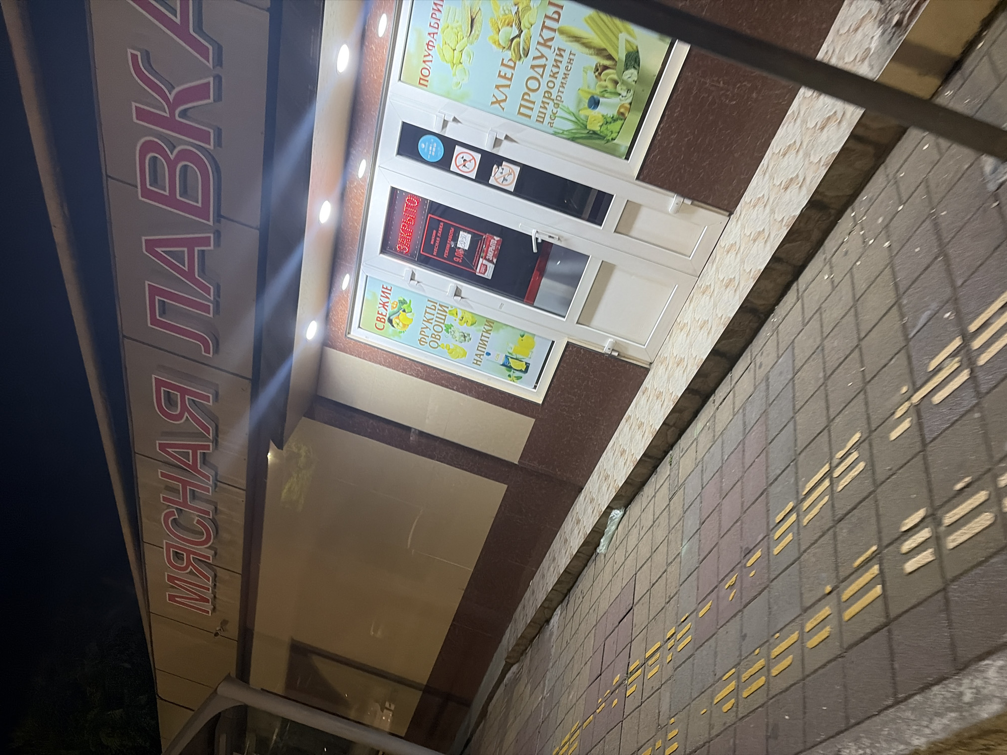 The image shows the facade of a store in Sochi, Russia, at night. The store's sign, written in red Cyrillic letters, reads "МЯСНАЯ ЛАВКА" (Meat Shop). Below the sign, there are windows displaying various products. One window advertises "СВЕЖИЕ ФРУКТЫ И ОВОЩИ" (Fresh Fruits and Vegetables) and "НАПИТКИ" (Drinks). Another window advertises "ПОЛУФАБРИКАТЫ" (Semi-finished products) and "ХЛЕБ И ПРОДУКТЫ ШИРОКИЙ АССОРТИМЕНТ" (Bread and Products, wide assortment). The entrance has two white doors, one of which has a sign indicating opening hours: 9:00-20:30, and it says "ЗАКРЫТО" (Closed). There are also two smaller signs with a prohibition symbol and a dog icon. The sidewalk in front of the store is paved with square tiles, some of which have yellow markings. The environment suggests an urban setting in Sochi.