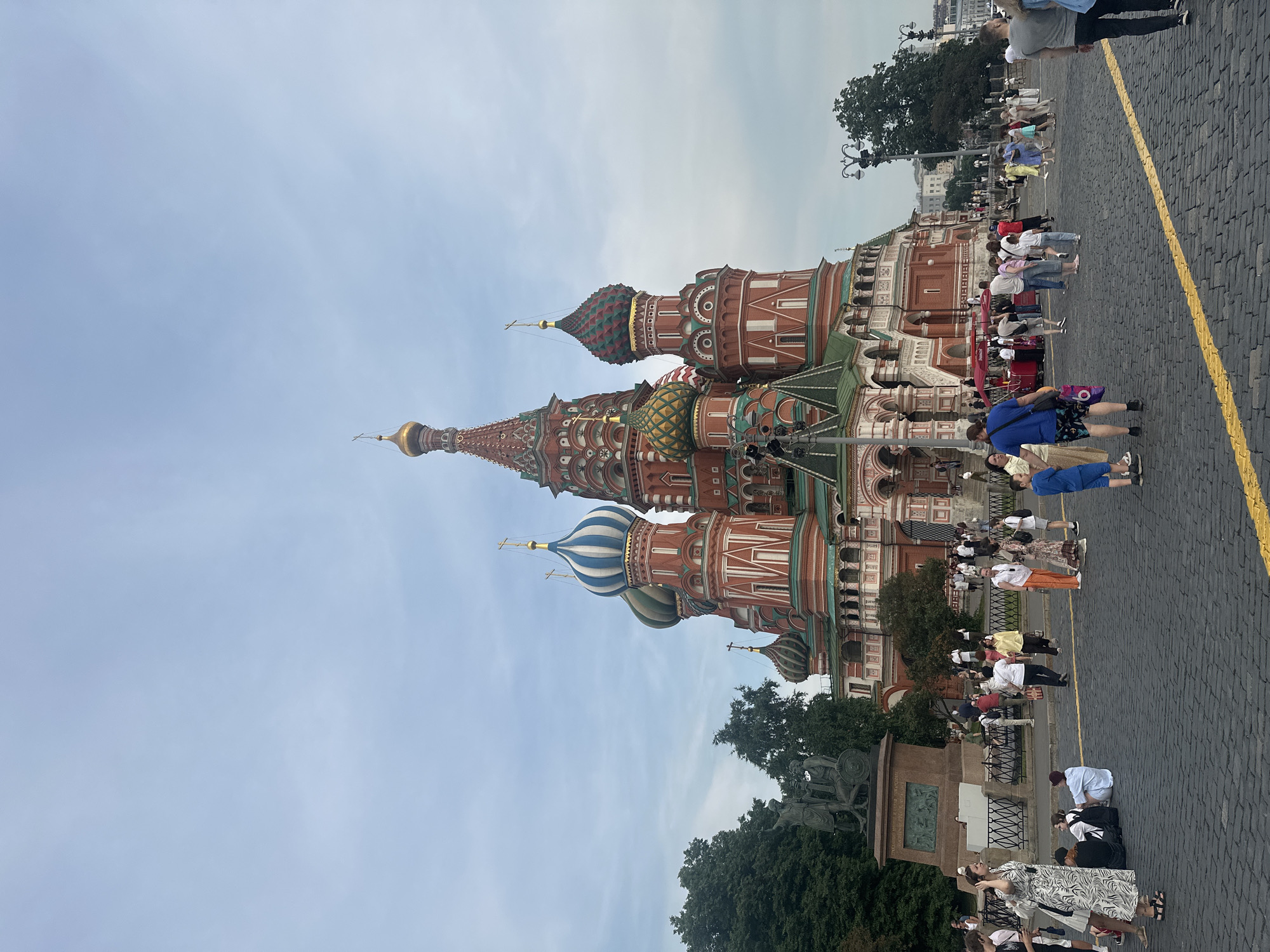 The image depicts Saint Basil's Cathedral in Moscow, Russia, a prominent landmark on Red Square. The cathedral's distinct onion domes, painted in vibrant reds, blues, greens, and gold, are the central focus, set against a pale blue sky with scattered clouds. Several individuals are visible in the foreground and midground, engaged in various activities. Some are walking, while others are seated on the cobblestone ground, likely tourists. A red tourist bus is parked nearby, and there is a statue in the background to the left of the cathedral, possibly a monument. The ground is paved with dark cobblestones, characteristic of Red Square, with a yellow line marking a pathway. Trees are visible on the left side of the image, and lampposts with streetlights are scattered throughout the scene. The overall impression is a bustling tourist area, with many people enjoying the architecture and atmosphere of this historic location.