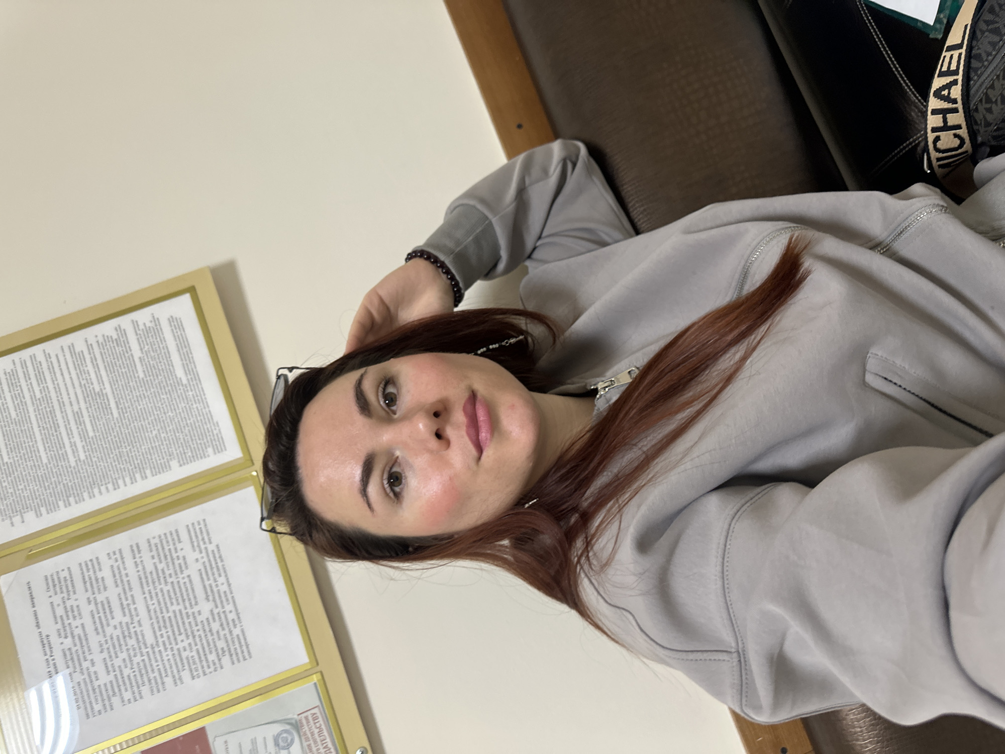The image shows a woman with long, auburn hair, wearing a gray hooded sweatshirt. She is leaning back on a brown leather couch. In the background is a beige wall with two framed documents. The woman is wearing a bracelet on her left wrist and has glasses tucked into her hair. A Michael Kors bag is visible beside her. The OCR text in the image identifies the location as Russia. The city of Mozhga is not explicitly mentioned in the text.