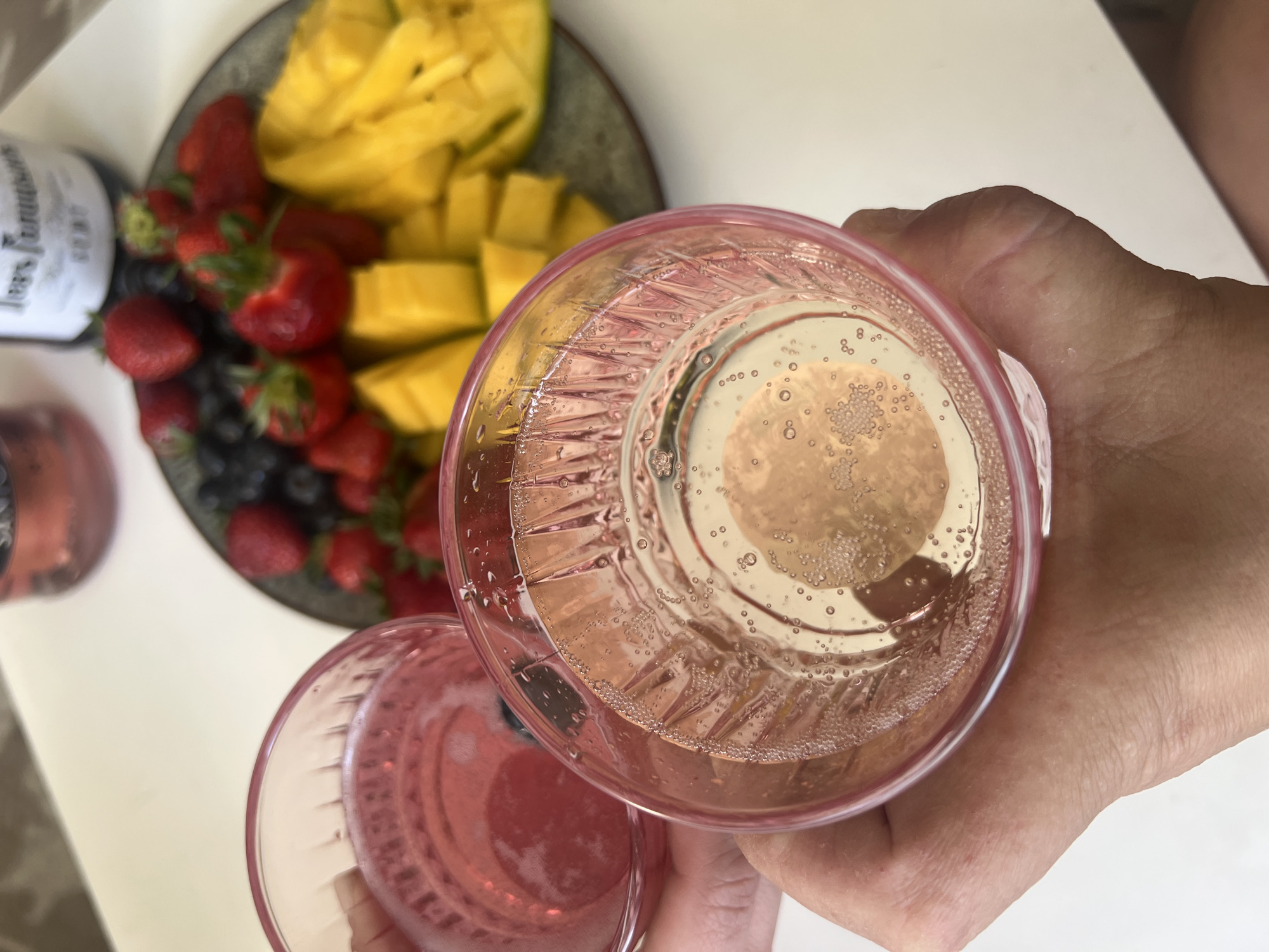 A person holds two glasses of sparkling wine above a plate of fruit. The plate contains chopped mango, strawberries, and blueberries. A bottle of sparkling wine is partially visible to the left. The background is a white surface. The location is not discernible from the image.