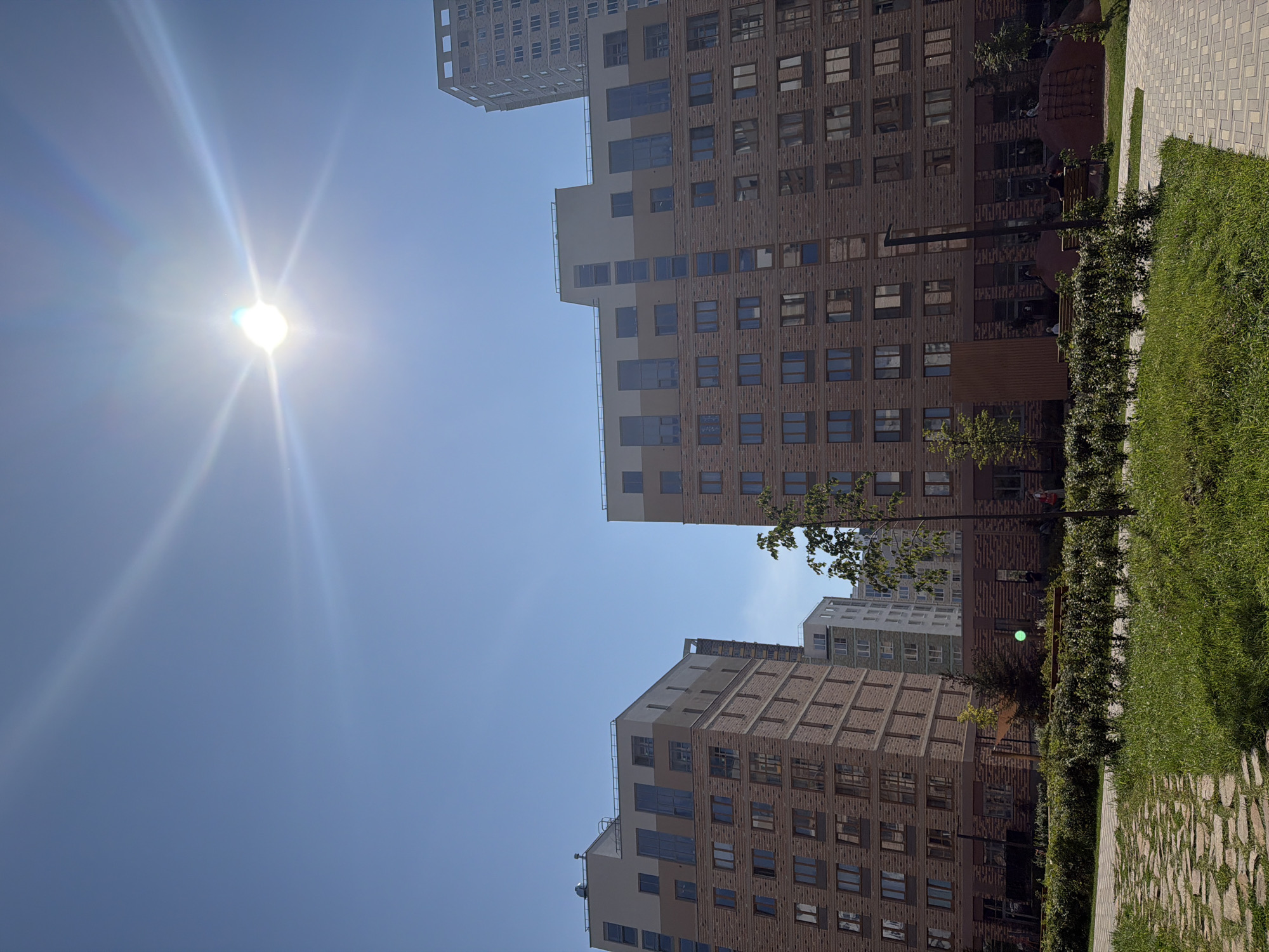 This is a full shot image of a modern residential complex in Yekaterinburg, Russia, on a sunny day. The sky is clear blue with the sun prominently visible at the top center, creating a lens flare. In the foreground, there is a paved pathway made of irregular stone slabs and a well-maintained green lawn with hedges. Two multi-story apartment buildings dominate the mid-ground. The building on the left is lighter in color with beige and brown brick facades, featuring numerous windows. The building on the right is similar in design but taller, with a facade primarily made of brown brick and a lighter beige upper section. Further in the background, another taller building with a white facade and visible window grids can be seen, along with other residential structures. A few trees and some low-lying greenery are scattered throughout the complex. In the lower part of the image, there are hints of people sitting on benches and a playground area.