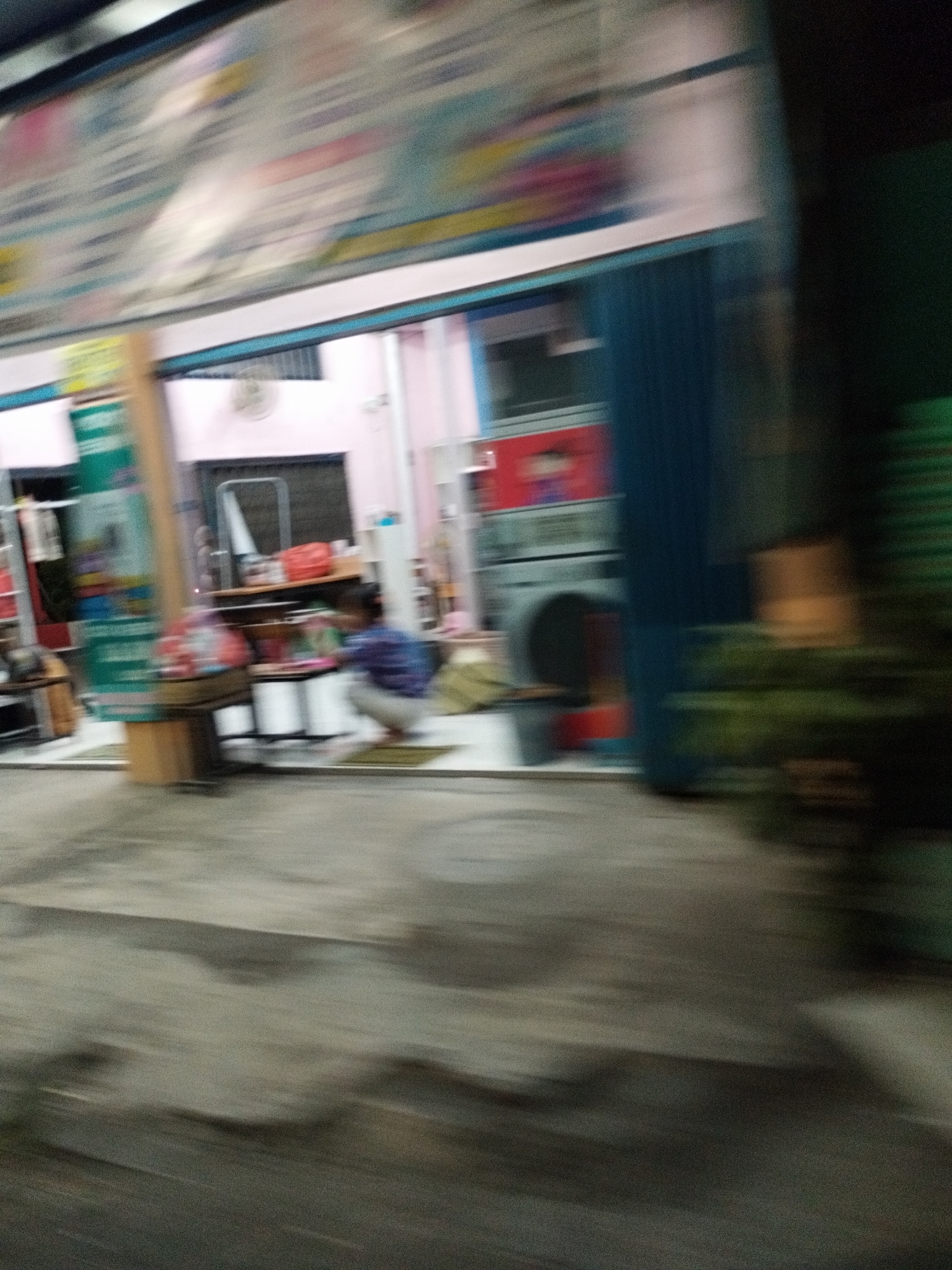 A blurred, motion-distorted image taken at night shows a storefront in Surabaya, Indonesia. The storefront features a glass-paneled entrance with pink-colored walls visible inside. To the left of the entrance, a green and white sign is mounted on a pole. Inside the store, shelves are visible, displaying various items. A person is seen crouching in front of the shelves. The ground in front of the store appears to be paved. The overall impression is of a commercial establishment with goods on display and a customer present.