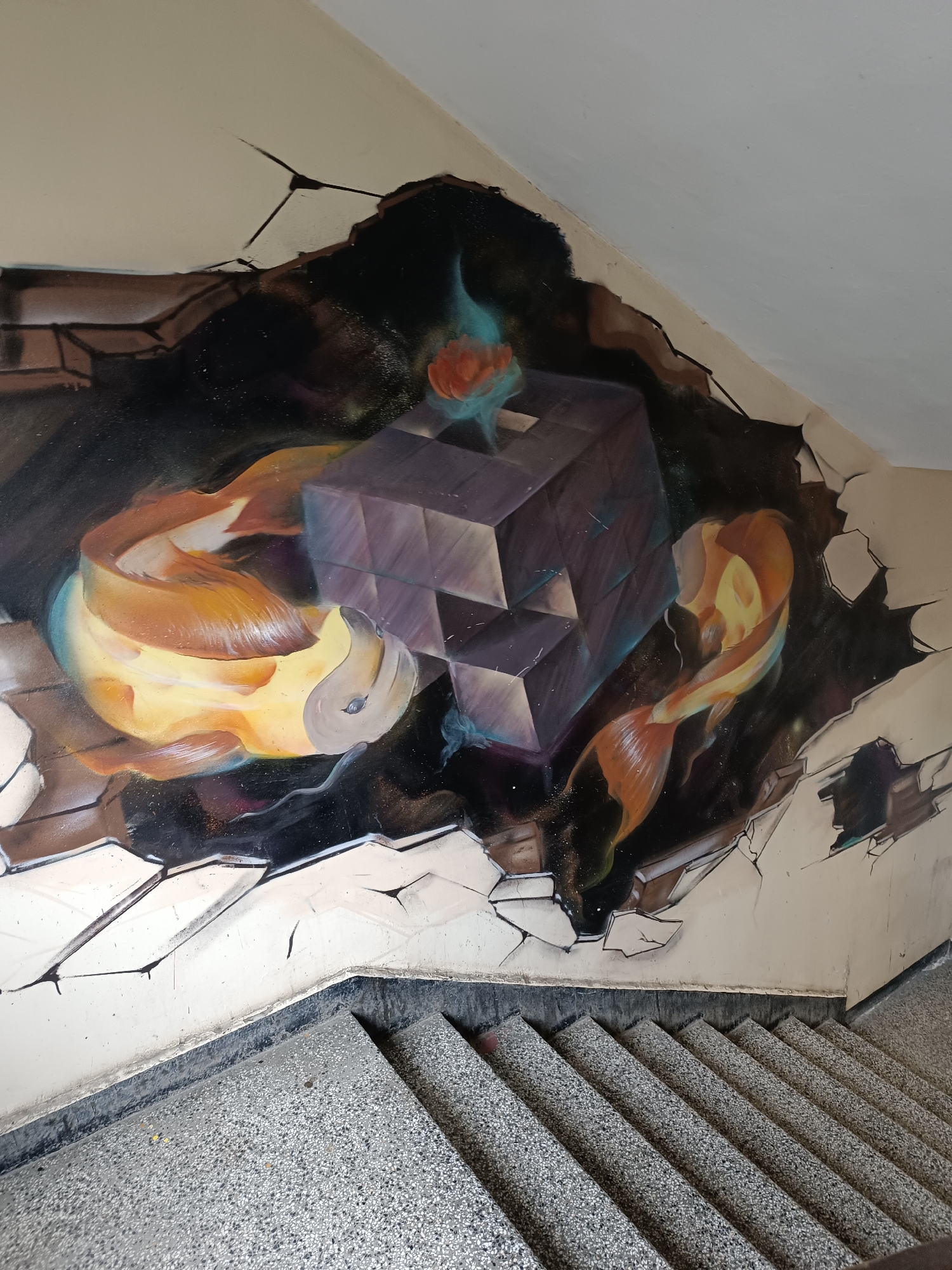This is a photograph of a staircase in Sofia, Bulgaria, featuring a mural on the wall. The mural creates an illusion of the wall breaking open to reveal a scene with two koi fish swimming around a geometric, cubic structure. The background beyond the "broken" wall is painted in dark colors, suggesting depth. The overall artistic style is reminiscent of graffiti art. The staircase is made of speckled gray concrete and leads downwards. The wall is painted a light cream color, which is visible around the mural.