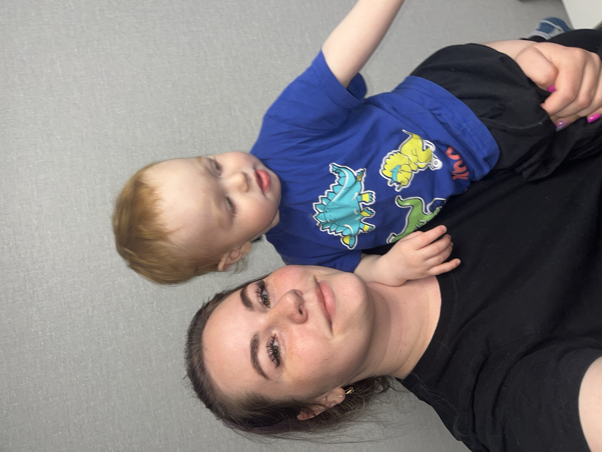A woman and a young child are in a selfie-style photograph. The woman is lying down, wearing a black shirt. The child is lying on top of her, wearing a blue shirt with cartoon dinosaurs on it. The background is a plain light gray wall. There is no visual indication of the location being in Chaikovsky, Russia.