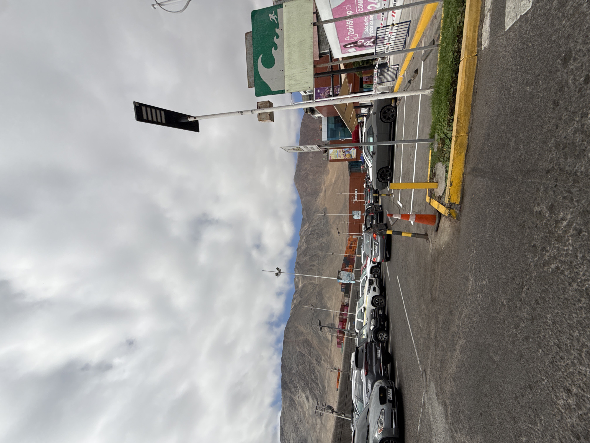 This image depicts a parking lot outside a commercial building in Iquique, Chile. The scene is dominated by a cloudy sky and a barren, mountainous landscape in the background. Several cars are parked in rows. A prominent feature is a tall pole with a modern LED street light at its top. Adjacent to it, a green sign with a white wave and a figure running is mounted on a pole. Further to the right, a commercial building with glass windows is visible, along with a sign for "ZofriShop.cl". The ground is asphalt, marked with parking lines and bordered by yellow curbs. An orange traffic cone is placed near a yellow bollard in the foreground. The overall impression is of a bright, somewhat overcast day in a coastal desert environment.