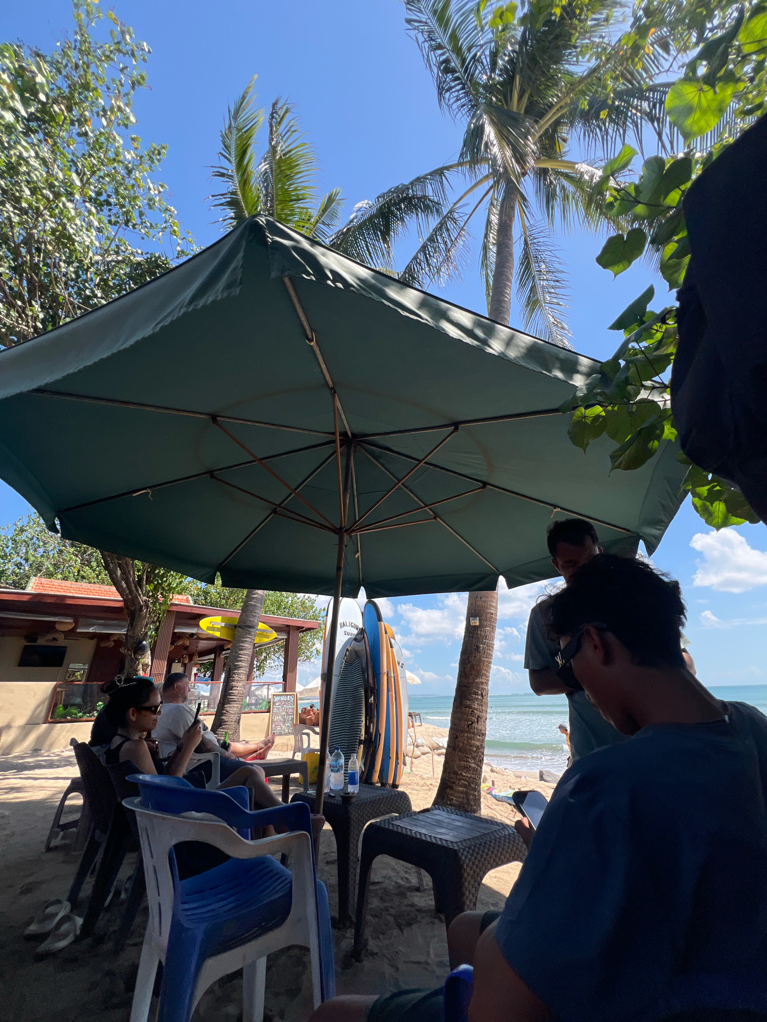 In Kuta, Indonesia, a beachside scene unfolds under a large green umbrella. The sandy ground is dotted with various chairs, some occupied by individuals. In the foreground, a person in a dark blue shirt faces away from the camera, looking at a mobile phone. To their left, a blue and white plastic chair is empty. Further back, a woman with dark hair and sunglasses is seated in a dark chair, also holding a phone. Beside her, a man in a white t-shirt sits in a white chair, looking at his phone. A surfboard is propped upright near a palm tree, alongside another surfboard with blue and yellow accents. In the background, a building with a red roof and windows is visible, possibly a restaurant or shop. Beyond that, the ocean stretches to the horizon under a clear blue sky with scattered clouds. Palm trees and other foliage frame the scene, with sunlight casting shadows on the sand.