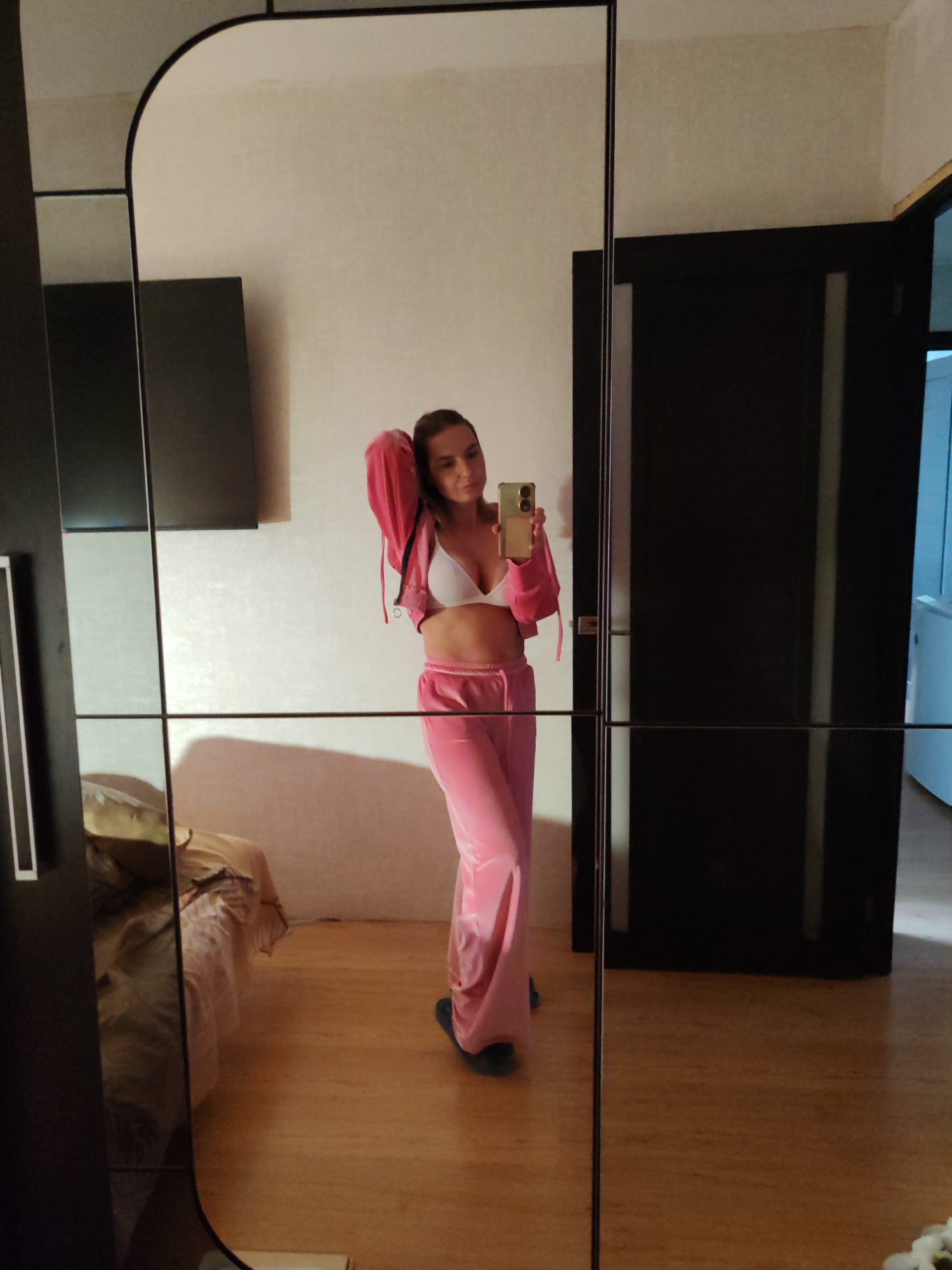 A woman in a pink tracksuit stands in front of a large, modern mirror that segments the reflection into panes. She is holding a smartphone, presumably taking a photo of herself. She is wearing a white bra and her pink jacket is slightly open. In the reflection, a bed and a flat-screen television can be seen. A closed dark door is to her right in the mirror. The floor appears to be wooden. The location is likely indoors, possibly a private residence. There is no verifiable evidence indicating the location is in Petropavlovsk-Kamchatsky, Russia.