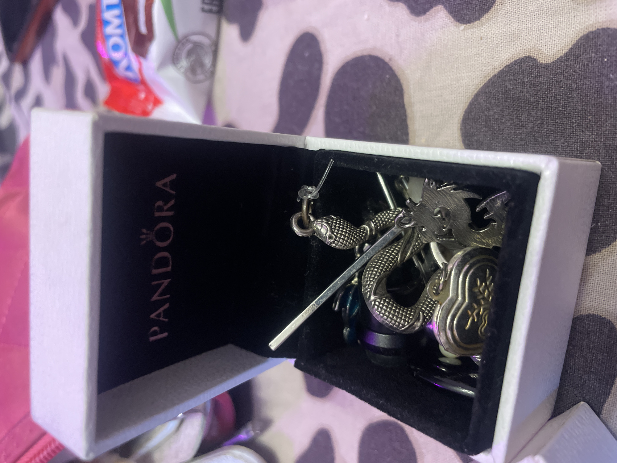 A white Pandora jewelry box is open and sits on a patterned surface. The inside of the box is lined with black velvet and contains several silver-colored charms. One charm is shaped like a snake wrapped around other charms. Another charm is in the shape of a heart with an intricate design. Also visible are a thin silver rod and some dark-colored beads. In the background, a red and white candy wrapper is visible. The scene appears to be indoors, possibly in a residential setting. There is no visible information to suggest this scene is located in Ladozhskoe Ozero, Russia.