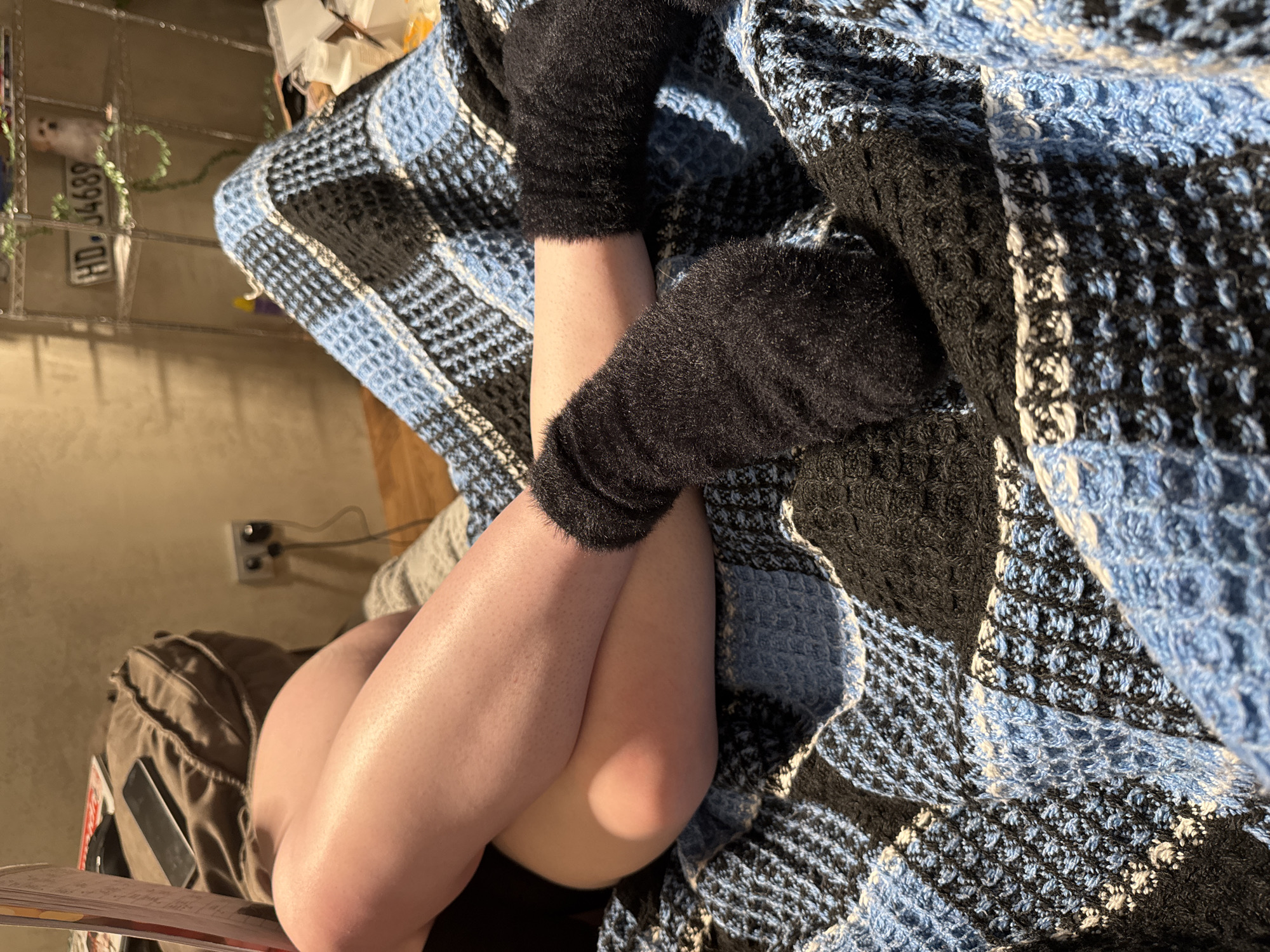 The image shows a close-up view of a person's legs and feet, partially covered by a blue and black checkered blanket. The person is wearing black fuzzy socks. One leg is crossed over the other. In the background, there is a blurry view of a bag, a phone, a wall outlet, and shelves with various items, including a small white dog figurine and a license plate with the numbers "HD.04689". The overall setting appears to be indoors, possibly a living room. The location is identified as Baravlyanski Sel'ski Savet, Belarus.
