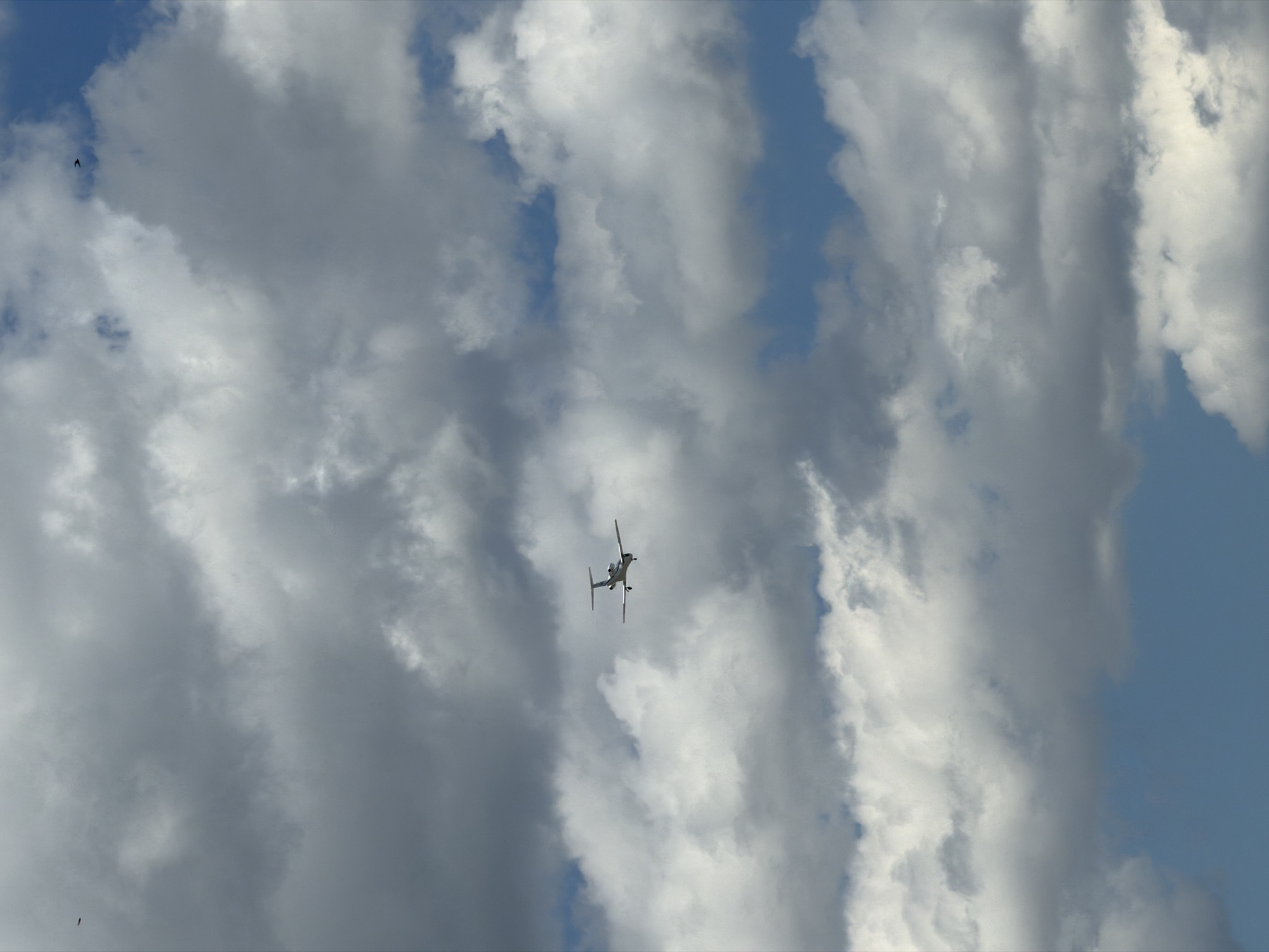 This media file captures a view of the sky, filled with dramatic, textured clouds. The scene is dominated by these large, white and gray cloud formations, interspersed with patches of bright blue sky. The lighting suggests it is daytime, with sunlight illuminating the edges of the clouds, creating a sense of depth and volume.
The primary object of interest is a small, light-colored aircraft, possibly a drone or a small plane, positioned in the upper center of the frame. It appears to be in flight, tilted slightly as if maneuvering. The aircraft is dark against the bright clouds, making it a distinct focal point. In the top left portion of the image, a very small, dark silhouette of a bird can be seen, adding another element of natural movement to the sky.
The overall mood is serene yet dynamic, due to the movement implied by the flying objects and the visual texture of the clouds. The vastness of the sky and the atmospheric conditions create a sense of openness and perhaps a touch of awe. There is no visible text in the image. The setting is outdoors, looking directly up into the sky.