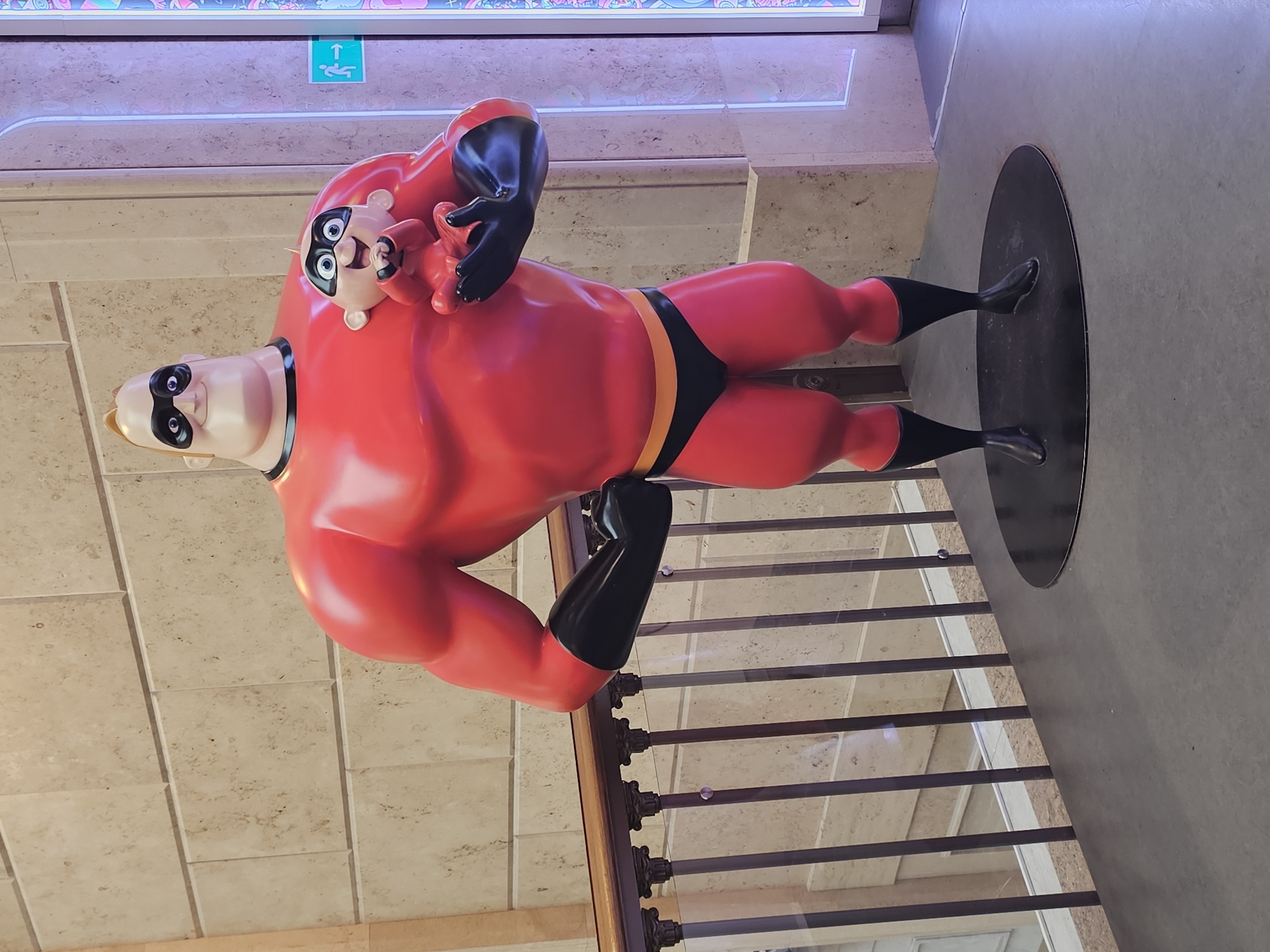 A life-sized statue of Mr. Incredible, from Disney's The Incredibles, stands prominently in an indoor setting. He is depicted in his iconic red and black superhero suit. His left arm is raised, cradling a smaller statue of Jack-Jack, also in his red outfit. The statues are positioned on a circular black base, which rests on a grey floor. Behind the statues, a dark brown railing with ornate pillars separates the area from a lower level. The background features a wall made of large, light-colored rectangular tiles. To the right of the statues, a brightly lit, decorative panel with pink and blue patterns is visible, along with a green exit sign with a running figure. The lighting in the scene appears to be a mix of ambient and artificial, with the decorative panel casting a colorful glow. The location is identified as Unknown, Unknown.