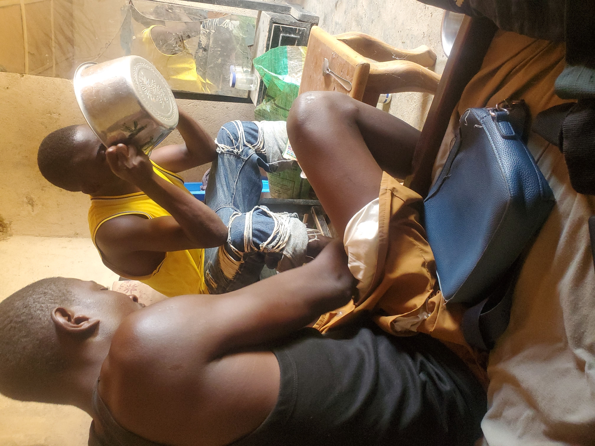 Two young men are seated on the floor in a sparsely furnished room in Wumat Central, Nigeria. The man in the foreground, viewed from the back, wears a black tank top. To his right, another man in a yellow tank top and ripped denim jeans is holding a large metal bowl to his face, appearing to drink or eat from it. A small wooden table with a spoon rests beside him. Various bags and containers are visible in the background, along with a metallic structure that may be a refrigerator or cabinet. The walls are rough and unpainted, suggesting a basic living environment. The overall impression is one of everyday life in a rural or underdeveloped urban setting.