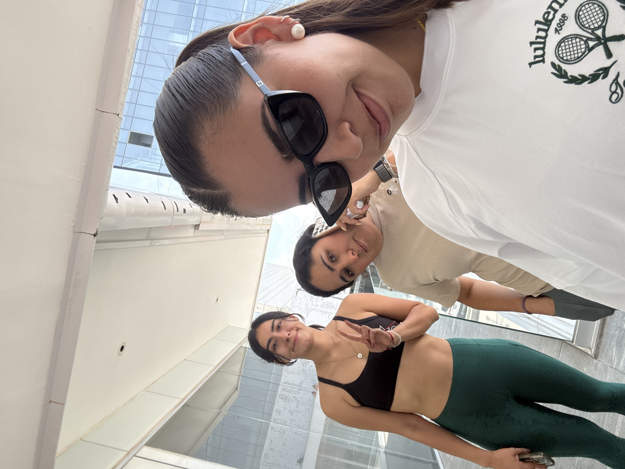 Three women are in the image. One woman is wearing sunglasses, a white shirt with a tennis emblem, and a watch. Another woman is wearing a beige shirt and is holding a phone. The third woman is wearing a black sports bra and green leggings, and making a peace sign with her hand. They are likely inside or on the edge of a building. The location is inferred to be Puebla, Mexico.