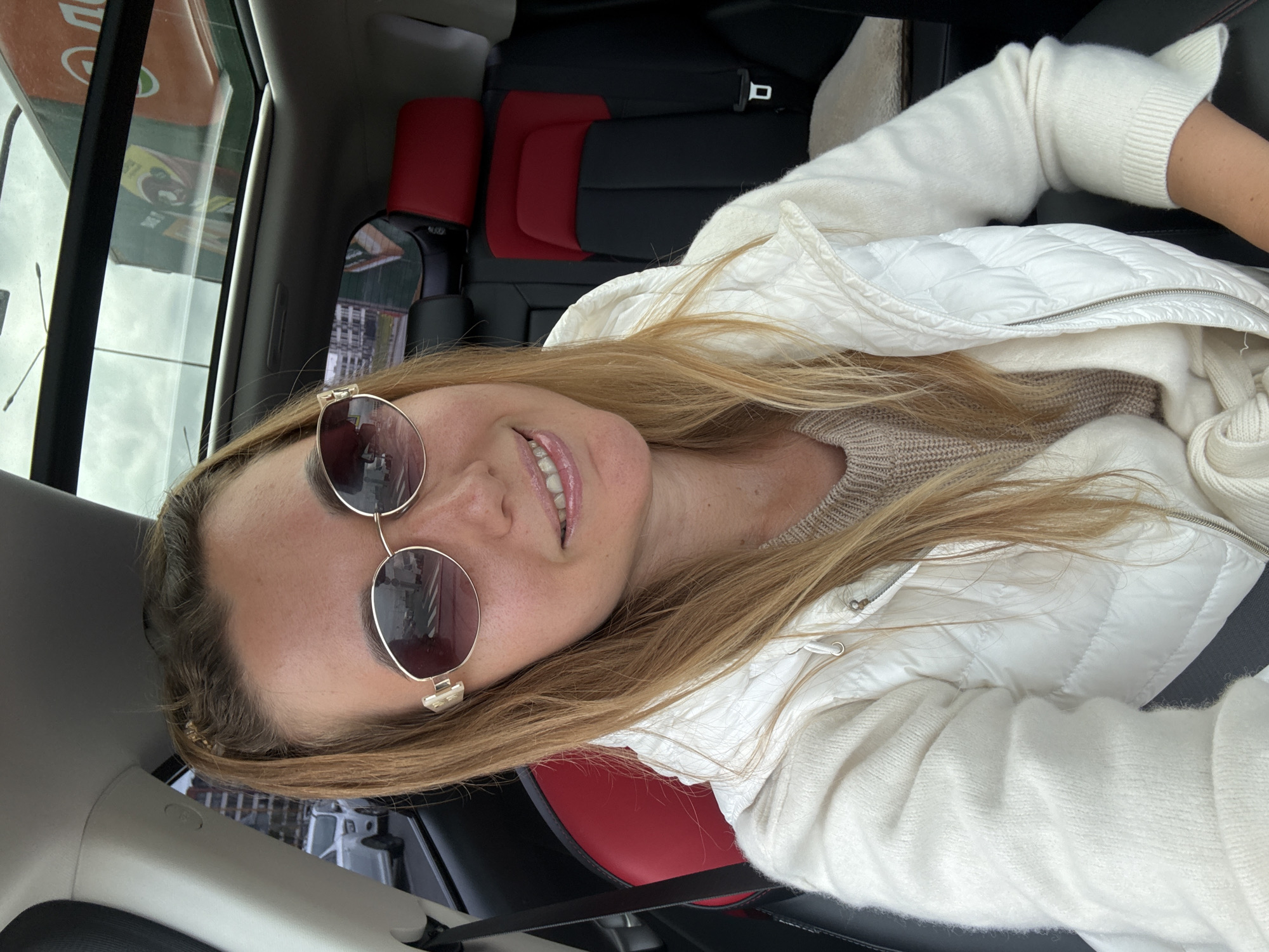 In this medium shot, a woman with long blonde hair, wearing sunglasses, a white puffer vest over a beige sweater, and a white scarf, is sitting in the passenger seat of a car. The car has a sunroof and black and red upholstered seats. She is looking at the camera and smiling. The background shows an urban environment with buildings and a cloudy sky visible through the car's windows.