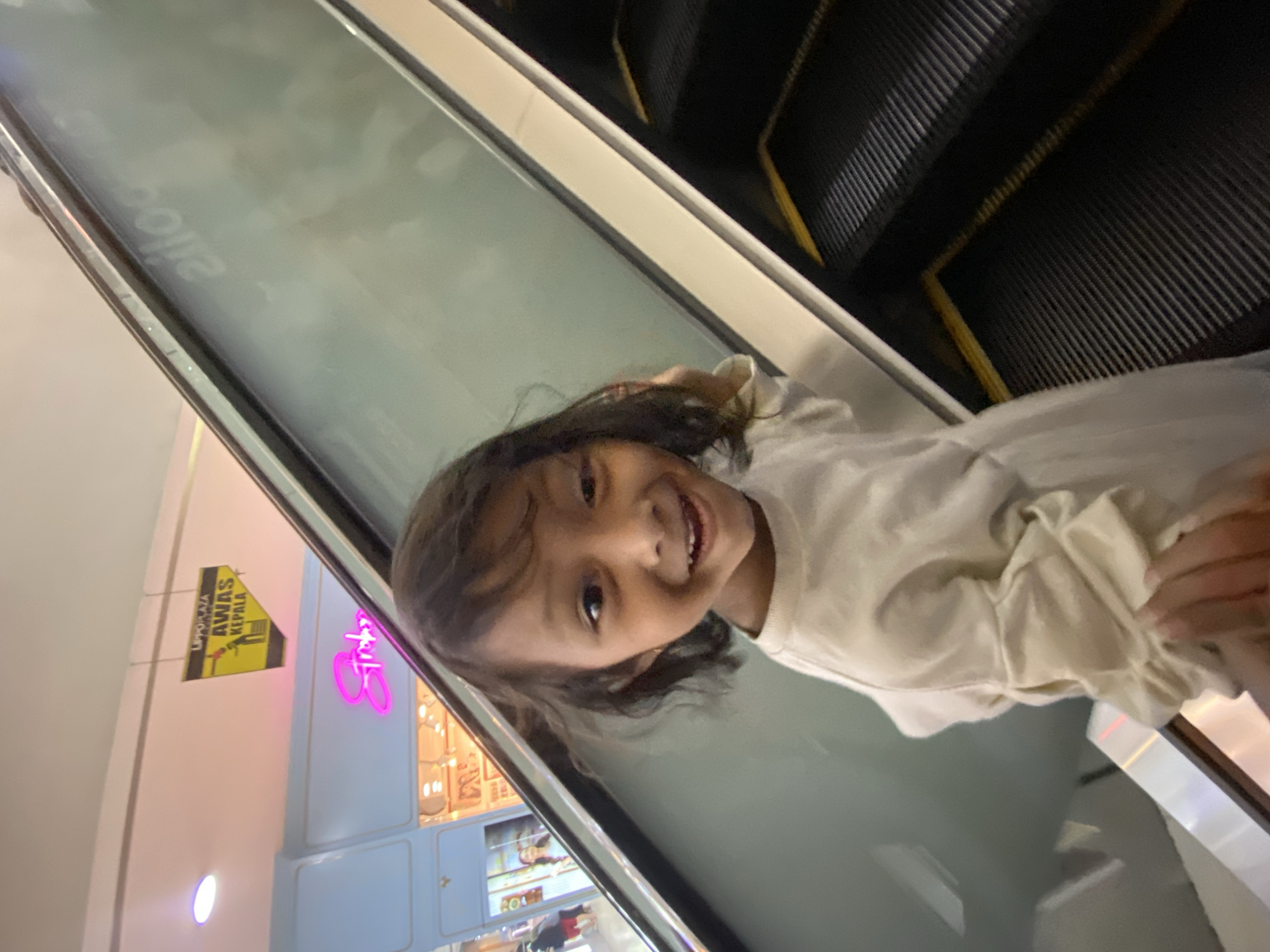 A young girl with dark, wavy hair and brown eyes smiles widely as she stands on an escalator. She is wearing a long-sleeved, cream-colored shirt. The escalator appears to be in a shopping mall, as indicated by the illuminated signage in the background, which includes "Lippo Plaza" and "AWAS KEPALA" (meaning "Beware of Head" in Indonesian), suggesting it is located in Sidoarjo, Indonesia. The girl is looking towards the camera, and her left hand is visible in the foreground, touching her shirt. The escalator steps are black with yellow safety lines.