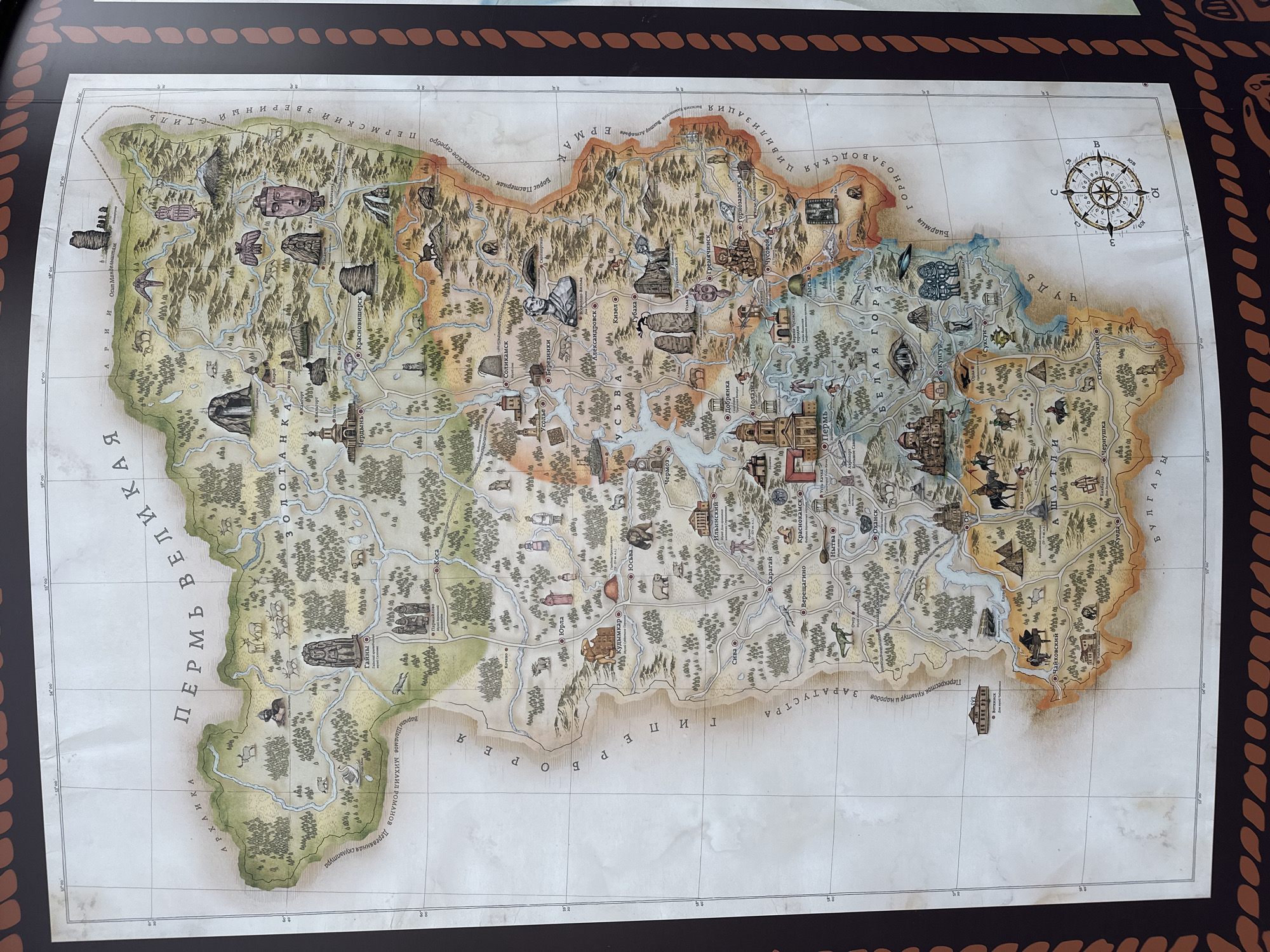 The image depicts a detailed, illustrative map. The map's content appears to be historical or stylized, showing geographic regions, settlements, and artistic renderings of various objects and figures. The map is not set in the city of Bolshoye Savino, Russia but rather covers a large area of Western Russia. The names of cities are visible such as Perm, Kudymkar, Cherdyn, Krasnovishersk, Solikamsk, Berezntki, and several other cities or villages. The map also has a compass in the upper-right corner and coordinates on the top-left and bottom-left. The map is mounted on a dark background, possibly a display or presentation surface. The environment suggests a museum or exhibition setting.