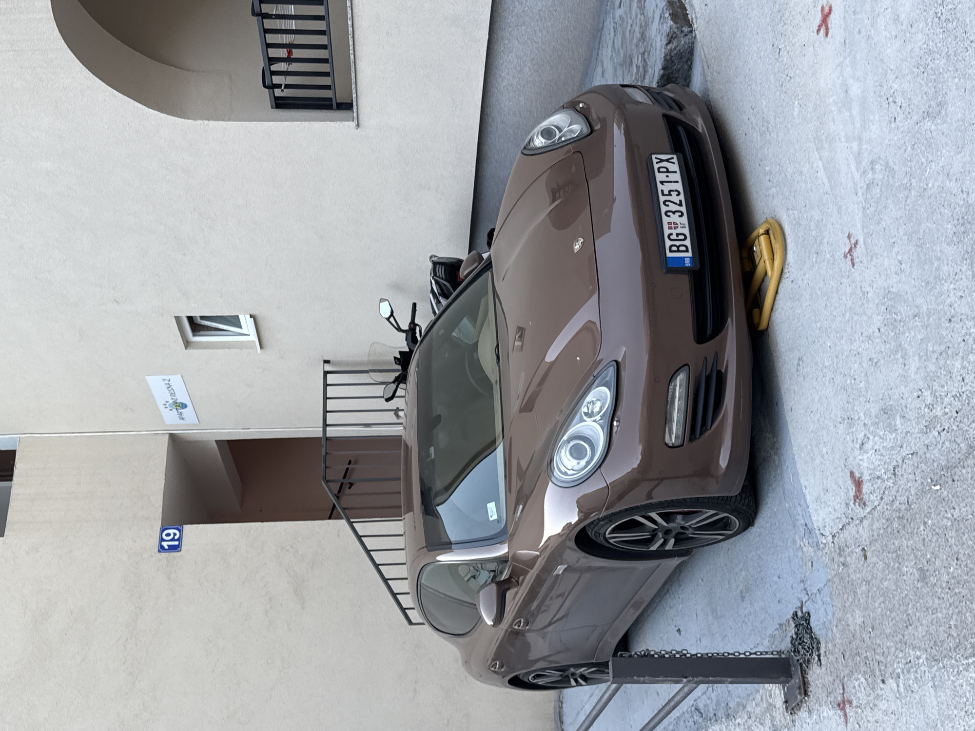 A brown Porsche Panamera is parked in an outdoor area, possibly a courtyard or alleyway, in Budva, Montenegro. The car has a yellow parking boot placed under its front wheels. A motorcycle is parked behind the Porsche, and a set of stairs with a black railing is visible to the left and behind the car. The building facade is light-colored, with an arched opening visible on the right side of the image. A sign with the number "19" in a blue rectangle is mounted on the wall near the stairs, and another sign with "APARTMANI RESNA 2" is also visible. The ground is concrete and appears somewhat weathered, with some red markings on it. The overall scene suggests a residential or commercial area in a European city.