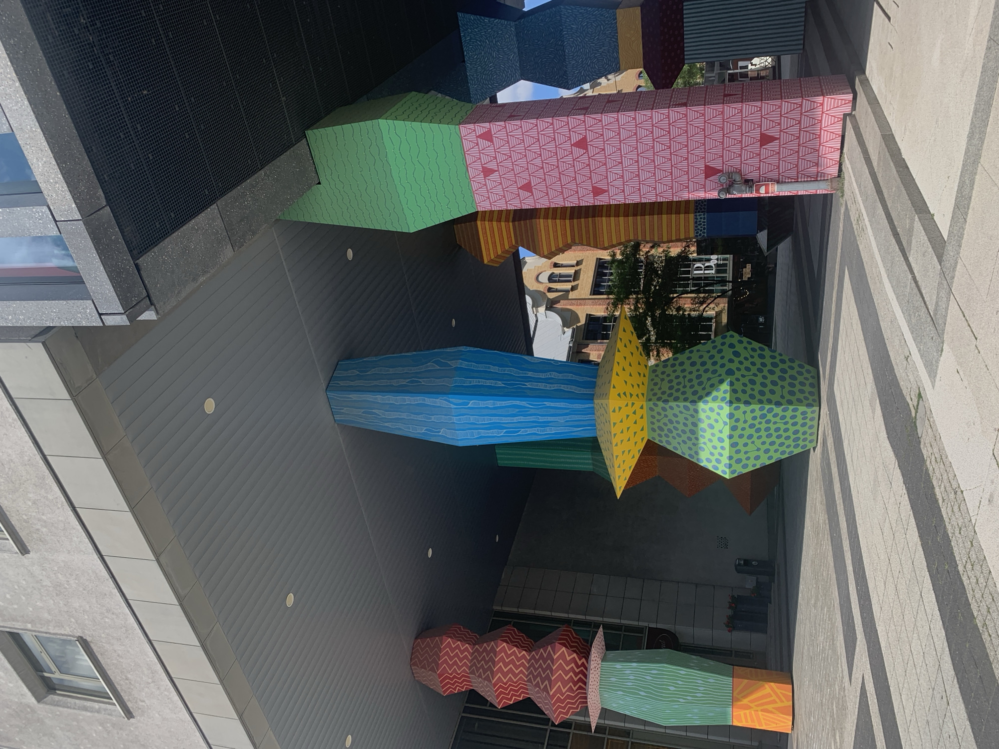 The image depicts an outdoor public space in Poznan, Poland, beneath a modern building with gray concrete and glass elements. The foreground is dominated by a series of large, colorful, polygonal sculptures that serve as decorative pillars. These pillars are adorned with various geometric patterns and vibrant colors, including green with white wavy lines and orange at the base of one, blue with white vertical lines, red with a zigzag pattern, yellow with triangular shapes, and pink with a repeating triangular pattern. The ground is paved with light-colored square tiles and darker rectangular accents, creating a striped effect where shadows fall from the sculptures and the building above. In the background, a brick building with arched windows and ornate architectural details is visible, suggesting a blend of modern and older city architecture. The sky is bright blue and visible through gaps in the overhang and between the sculptures. No people are present in the image.