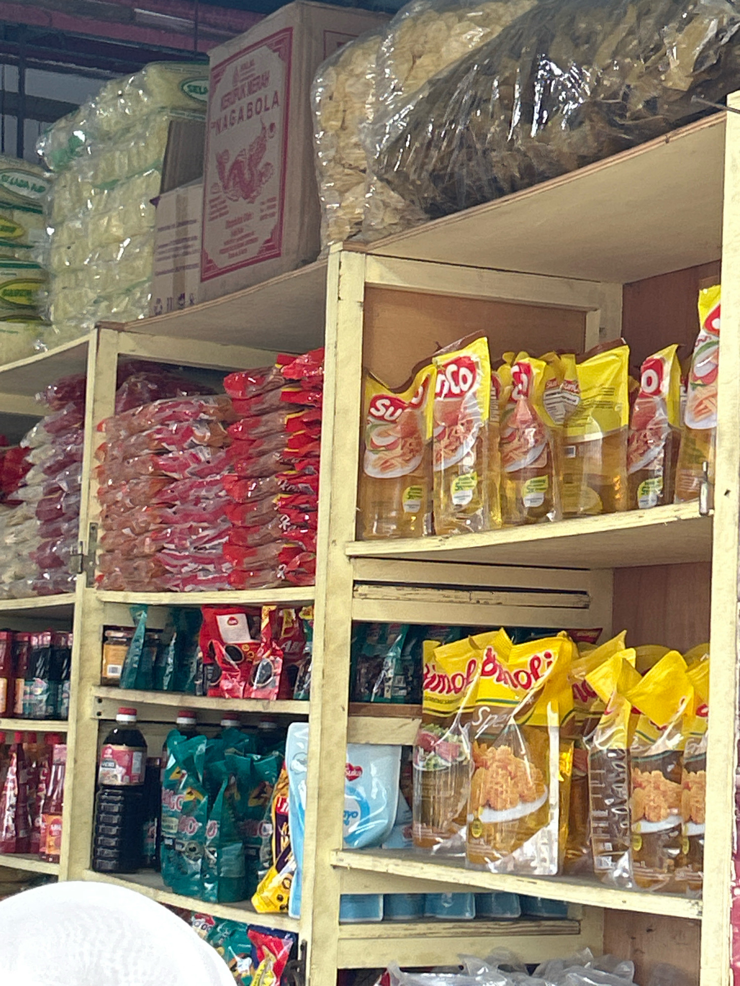 The image displays shelves stocked with various food products in a store, likely a traditional market or a small grocery store in Banda Aceh, Indonesia. The shelves are made of wood and are filled with packaged goods.
On the top shelf, on the left, stacks of items wrapped in clear plastic are visible, some with green labels. To the right of these, a large cardboard box with red and white labeling, featuring the text "KERUPUK MERAH NAGABOLA" and an illustration, is prominently displayed. Further to the right, a large, clear plastic bag filled with a yellowish, dry food product, possibly chips or crackers, is present.
The middle shelf is divided into two sections by a wooden divider. The left section contains numerous small red and pink packages stacked horizontally. The right section showcases several bright yellow pouches of cooking oil, with the brand name "SICO" visible on them. These pouches have images of food and nutritional information printed on them.
The bottom shelf holds a variety of items. On the left, a tall dark bottle with a red cap, likely soy sauce, is placed. Next to it are several green pouches, also packed with food products, and a blue and white container. To the right, more yellow pouches similar to those on the shelf above are visible. Below these, on a lower shelf, further packaged goods are visible, including some that appear to be snacks in clear plastic bags.
The overall impression is that of a well-stocked retail space, characteristic of food vendors or small supermarkets in urban areas of Indonesia, and specifically within the city of Banda Aceh, given the implied context. The lighting suggests an indoor environment, possibly with overhead fluorescent lights or natural light from an unseen source.