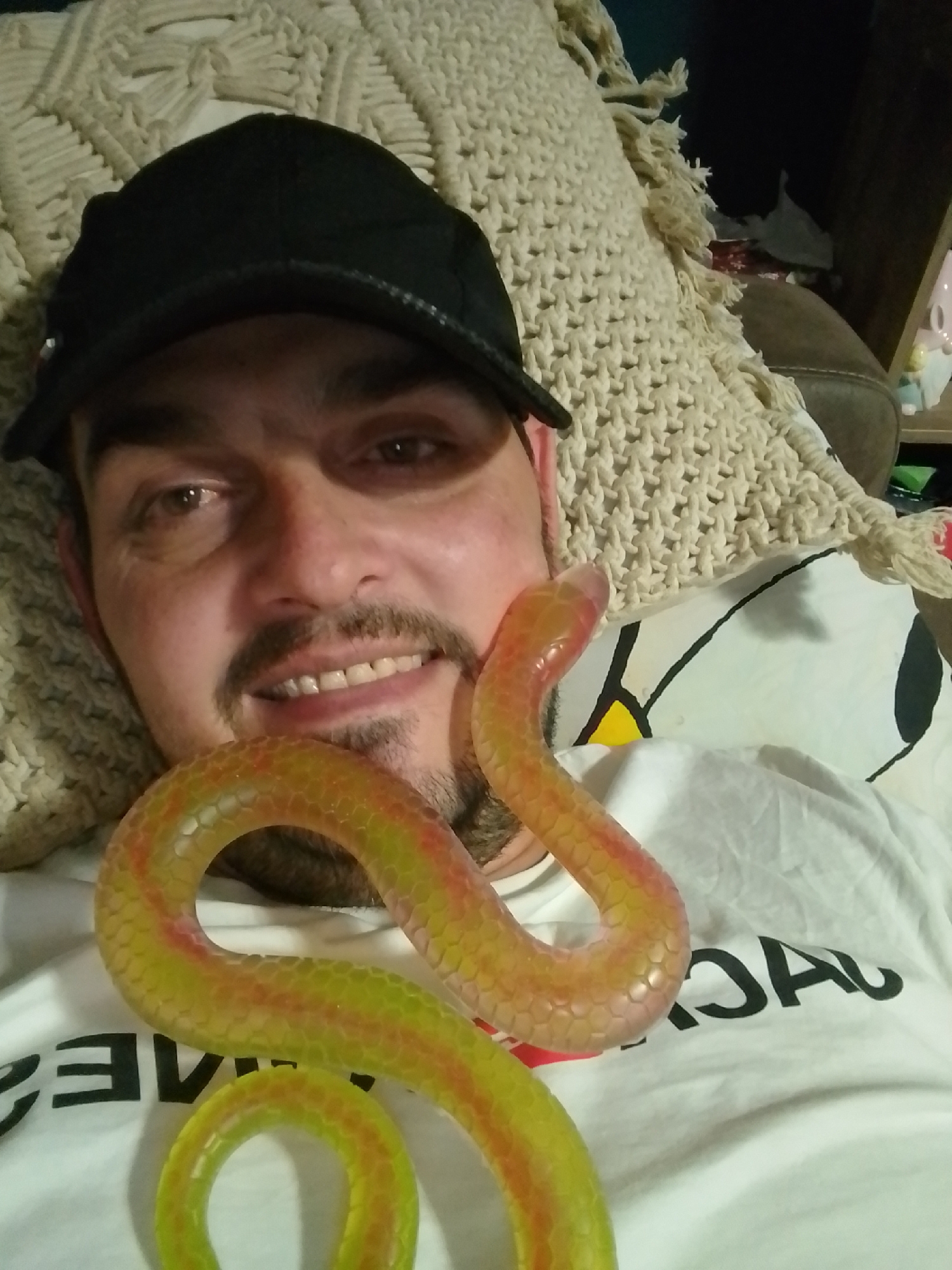 The image shows a man wearing a black baseball cap and a white t-shirt with black lettering. He is lying down, with a knitted blanket or throw behind his head. A translucent, snake-shaped toy is draped across his chest and neck. The man has a mustache and appears to be smiling. The lighting suggests an indoor setting. The image does not provide any information about the city of Wedel, Germany.