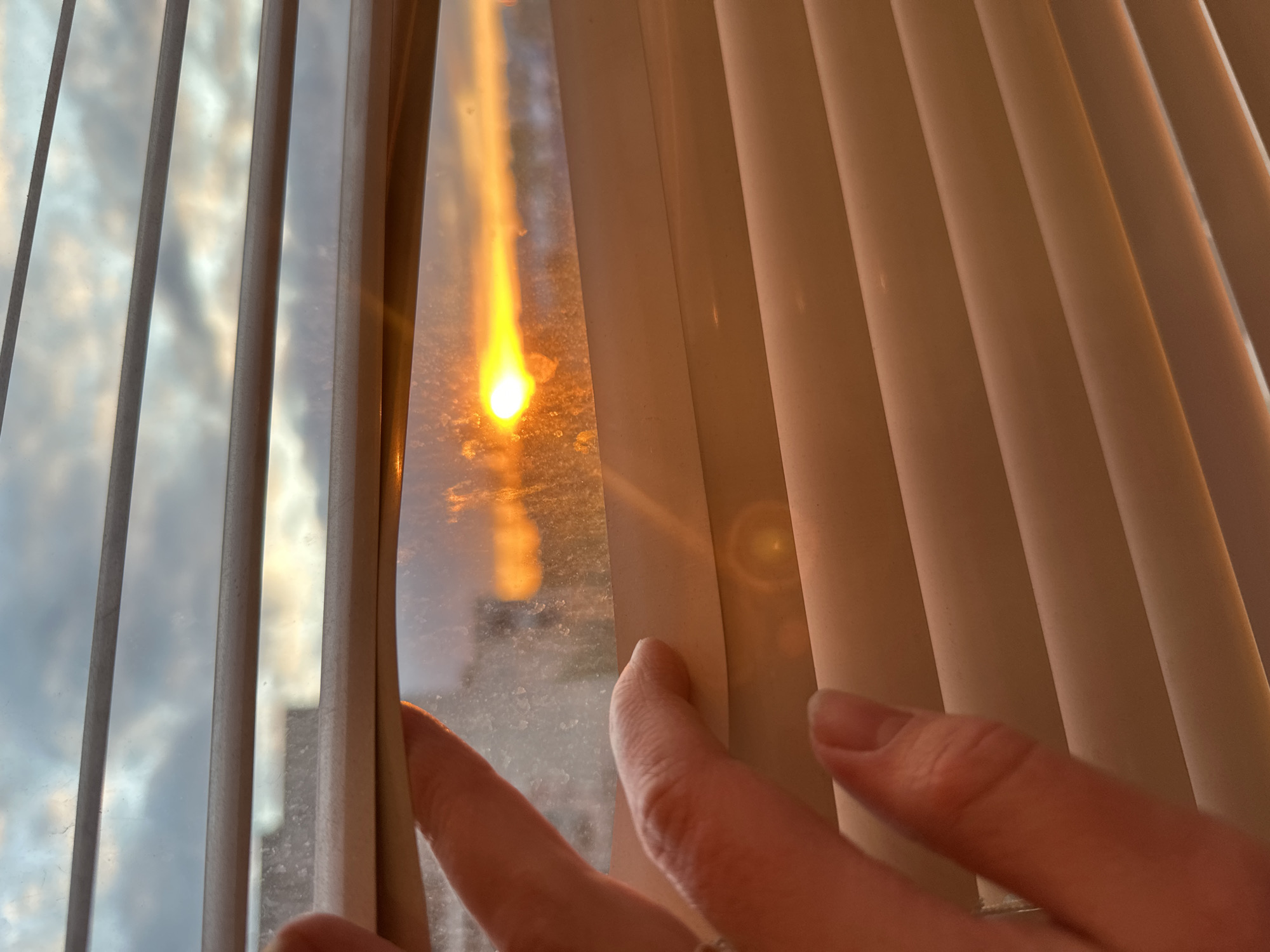 An indoor shot captures a hand with fingers gently pushing aside vertical window blinds to reveal the outside world. The scene is illuminated by the warm, intense glow of the sun, which appears as a bright, fiery orange and yellow orb in the upper left portion of the windowpane, casting a long, vertical streak of light. This suggests either sunrise or sunset.
Through the slightly dirty or dusty window, a cloudy sky with varying shades of grey and white is visible beyond the blinds, reflecting diffuse light. The blinds themselves are light-colored, likely off-white or beige, and are made of a semi-rigid material, curving gently where the hand separates them. A circular lens flare is discernible on the right side, within the shadow cast by the blinds, hinting at the direct angle of the sun. The hand, with manicured fingers and a ring on one finger, is positioned on the lower right, actively pulling back the blind slats to offer a peek. No visible text is present in the image. The location context is Tomilino, Russia.