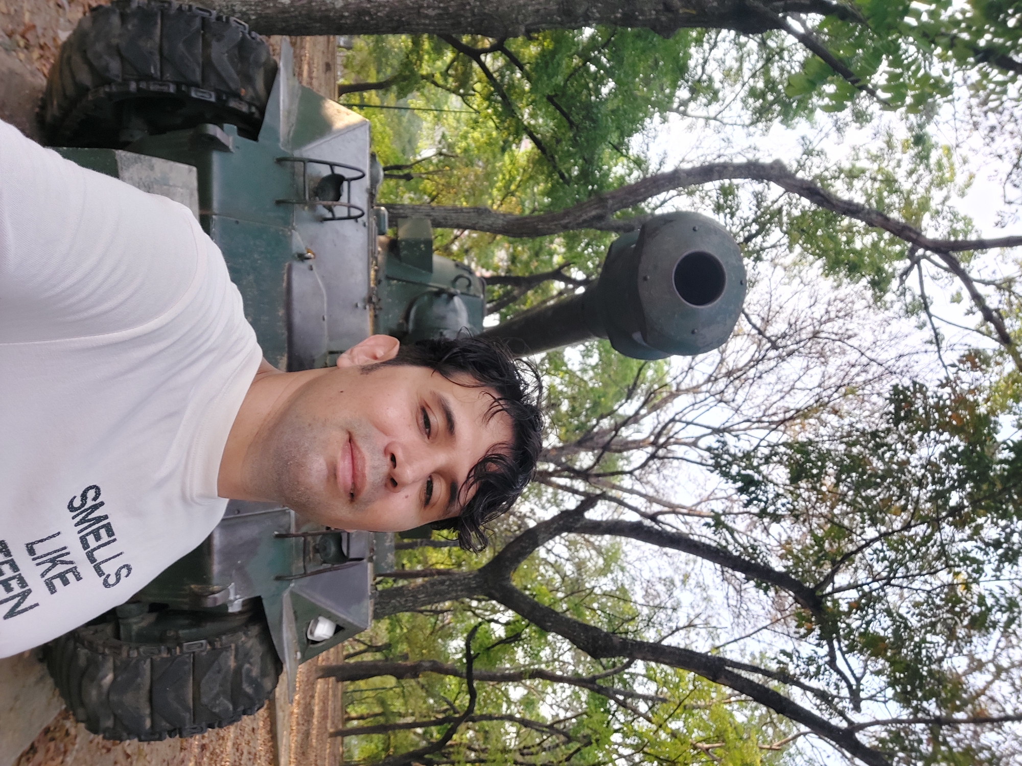 A man is pictured in front of a military tank at Parque del Este, Caracas, Venezuela. The man, wearing a white shirt, is taking a selfie. Behind him is a green military tank. In the background are trees and foliage.