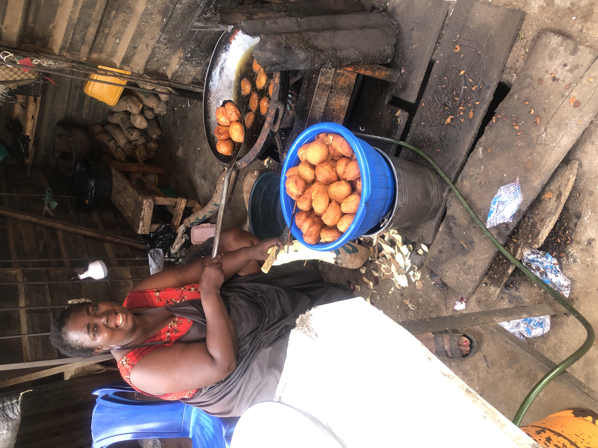 This image captures a vibrant, informal food stall scene, likely in Onitsha, Nigeria, given the provided context. The primary subject is a smiling woman, presumably the vendor, who is engaged in preparing and selling fried dough balls. She is seated in the foreground on a blue plastic chair, wearing a red patterned top and a dark wrapper/skirt, with her toenails painted red. She holds a small knife or peeler in her left hand.
To her immediate right, a large metal frying pan sits over a makeshift heat source, with several golden-brown dough balls sizzling in hot oil. A long-handled metal spoon, laden with more fried dough balls, is poised to remove them from the pan. Next to the frying setup, a blue plastic sieve or basket overflows with freshly fried dough balls, indicating a brisk trade.
The setting is rustic and functional, suggesting an outdoor or semi-sheltered market environment. In the background, a pile of yams or similar tubers is visible, alongside a yellow plastic basin and other miscellaneous items under what appears to be a corrugated iron roof. The ground around the stall is earthen and worn, with some discarded food scraps and plastic bags, indicative of a busy, lived-in space. A green hose is visible snaking across the ground.
The time of day appears to be daytime, with general bright but diffused lighting. The woman's warm smile creates a friendly and inviting atmosphere. There is no discernible text visible in the image.