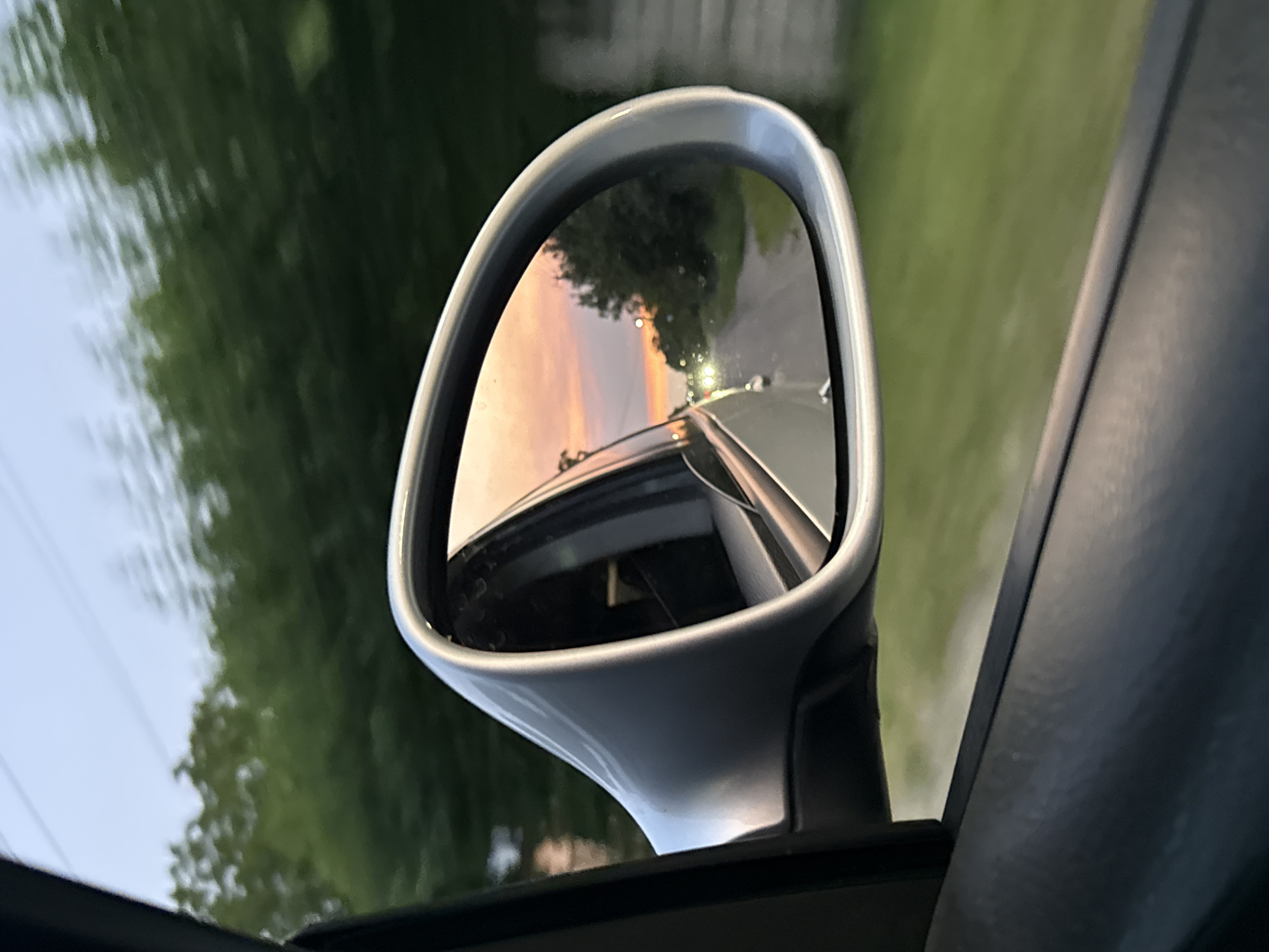 This media file captures a reflection in a car's side-view mirror, looking backward from the perspective of a moving vehicle. The mirror, a sleek silver color, is a prominent object in the foreground. Within the mirror's reflection, a sunset sky is visible, painted with hues of orange, pink, and yellow. This warm sunset contrasts with the cooler tones of the sky on the left side of the overall image.
The reflection also shows a road stretching into the distance, illuminated by streetlights. The road appears to be asphalt, and there are hints of other vehicles or light sources further down. Trees with green foliage line the sides of the road, blurred due to the motion of the car. In the background of the overall image, outside the mirror, the same blurred green trees and a sliver of pale sky are visible.
The time of day is clearly sunset, as indicated by the sky's colors and the streetlights becoming illuminated. The weather appears clear and calm, suitable for driving. The scene suggests a journey through a semi-rural or suburban area, likely in Petrushky, Ukraine, given the location context. There is a sense of movement and perhaps a moment of peaceful observation during travel. No text is visible in the media file.