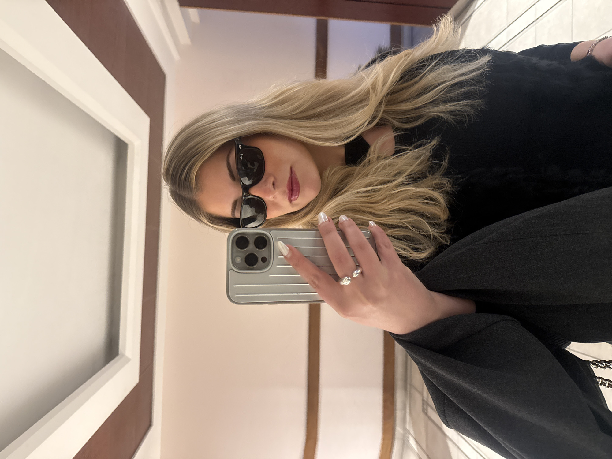 A young woman with long, wavy blonde hair is captured in a mirror selfie, rotated 90 degrees clockwise. She wears dark, rectangular sunglasses, which appear to be Ray-Ban from the faint branding on the arm, and a glossy reddish-pink lipstick. Her outfit includes a dark, possibly black, top or jacket with some texture, and dark grey wide-leg trousers.
She is holding a light grey smartphone, likely an iPhone given its triple-camera array and distinctive ridged case, with her right hand. Her fingernails are painted white or an iridescent color, and she wears two silver-toned rings on her right index and middle fingers.
The setting is indoors, reflected in the mirror. The background shows a well-lit room with architectural features, including a prominent white frame bordering a section of what appears to be a light brown or wooden wall panel. The overall scene suggests a stylish and casual moment. There are no specific visual cues within the image itself to indicate the provided context of Paris, France. No visible text apart from the faint branding on the sunglasses.