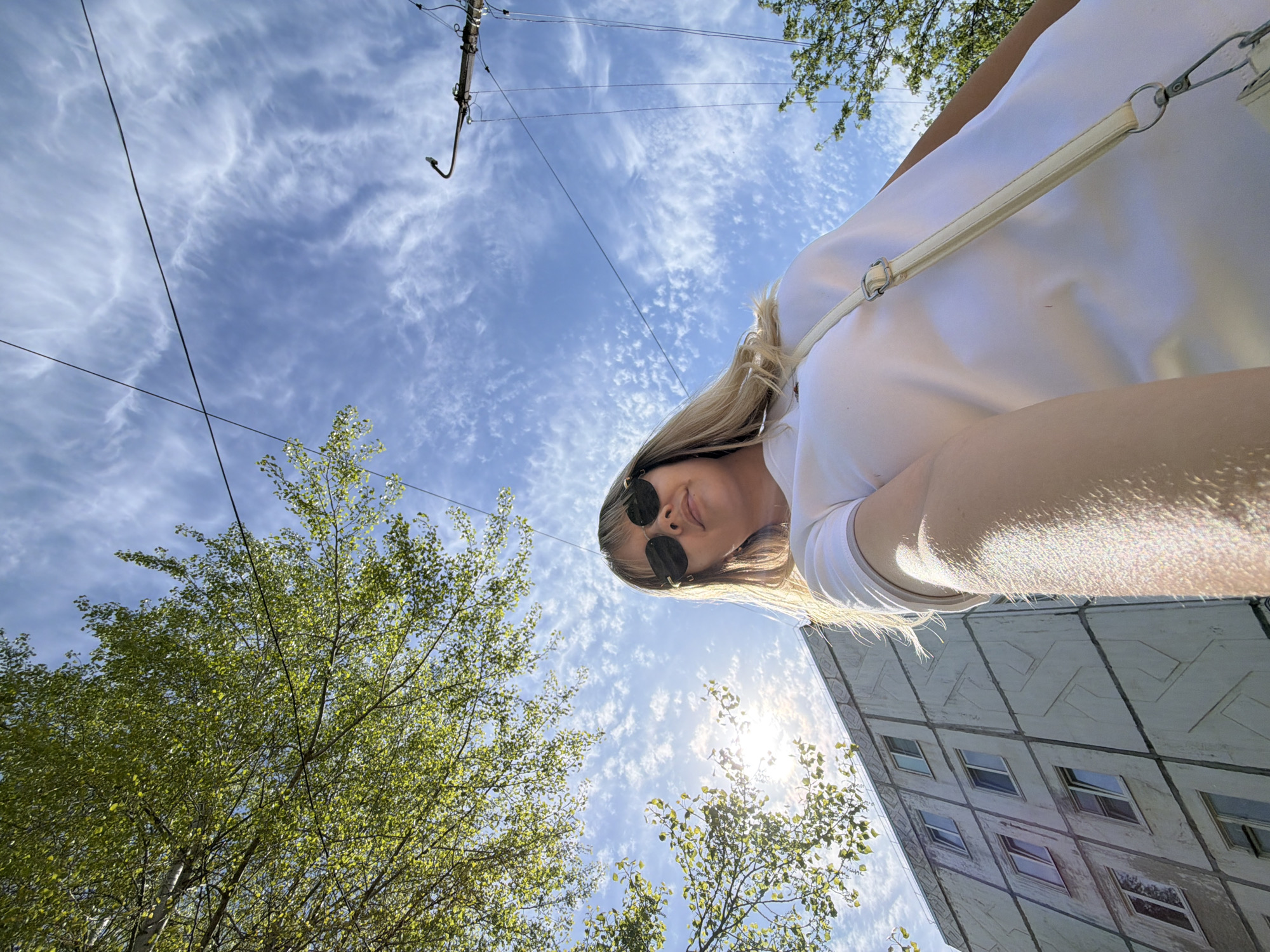 A low-angle, upward-pointing photograph shows a blonde woman with sunglasses, wearing a white top with a white strap slung across her chest, standing somewhere in the city of Chaikovsky, Russia. The sky above is blue with wispy clouds, crisscrossed by power lines. Trees with light green foliage appear on the left and bottom edges of the frame. To the right of the woman and slightly below her, part of a multi-story building is visible, constructed with large, geometrically patterned concrete panels. Sunlight is visible through the branches of the trees in the lower center of the frame.