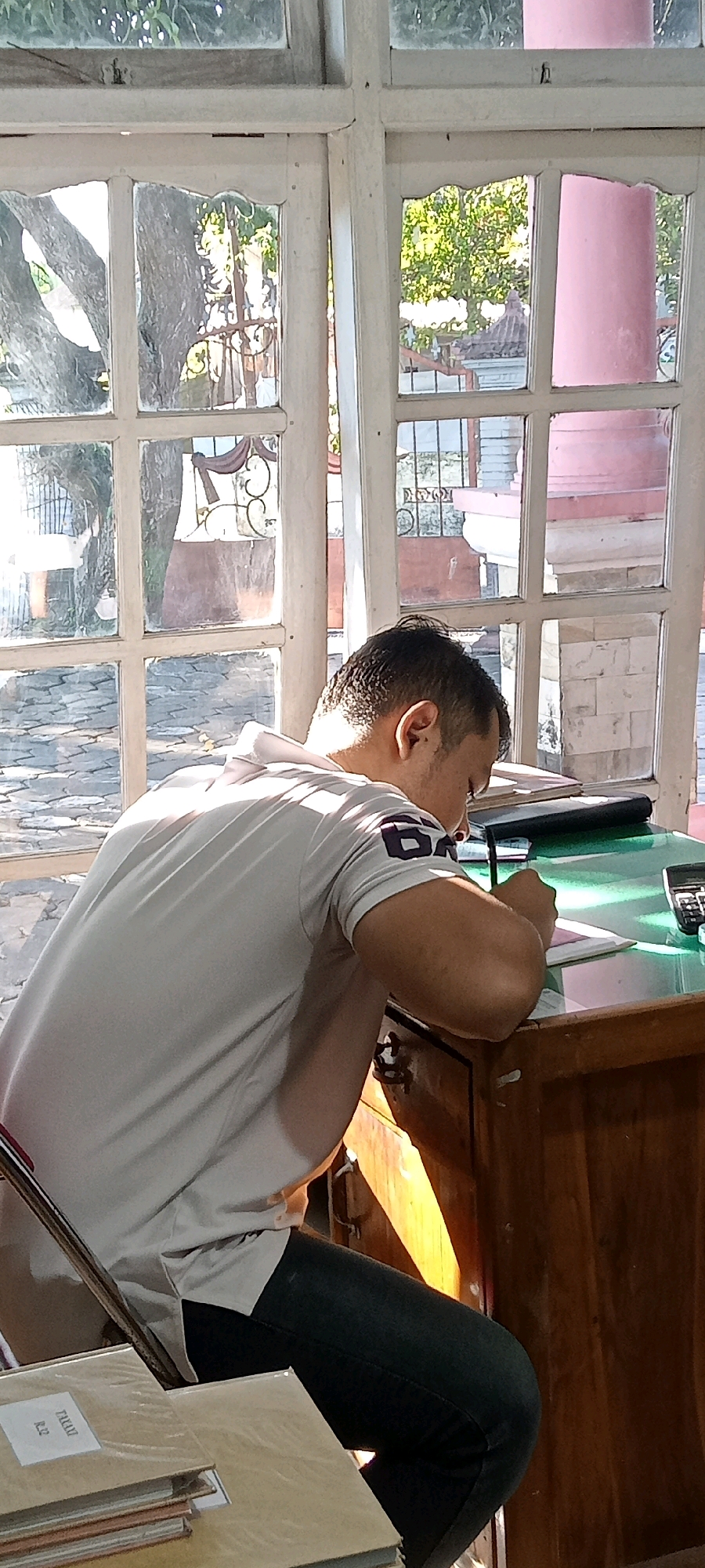 A man is sitting at a wooden desk, writing in a notebook. He is wearing a light-colored t-shirt with a dark logo on the sleeve and dark pants. The desk has a green surface and a calculator is visible on the right side. In the background, there is a large window with multiple panes, through which trees and buildings are visible. The scene appears to be indoors, possibly in an office or study. The light coming from the window suggests it is daytime. The man is focused on his writing, with his head bent down over the notebook. The overall impression is one of quiet concentration. This scene is in a building in the city of Begajah, Indonesia.
