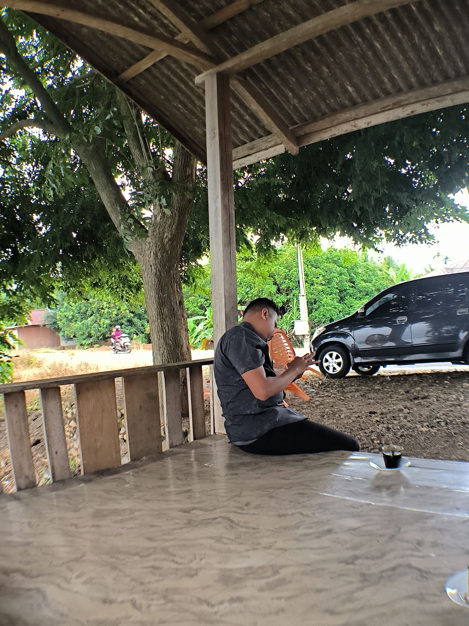 A man is seated on a concrete surface under a covered wooden structure. He is wearing a dark-colored short-sleeved shirt and black pants and is looking down at his phone. An orange plastic chair is beside him. In the background, a dark-colored car is parked on a gravel surface. Further back, trees and vegetation are visible, with a glimpse of a building and a person riding a scooter. A small cup of dark liquid sits on a saucer to the man's right. The scene appears to be outdoors, likely in a rural or semi-rural setting in Teupinreusep, Indonesia.