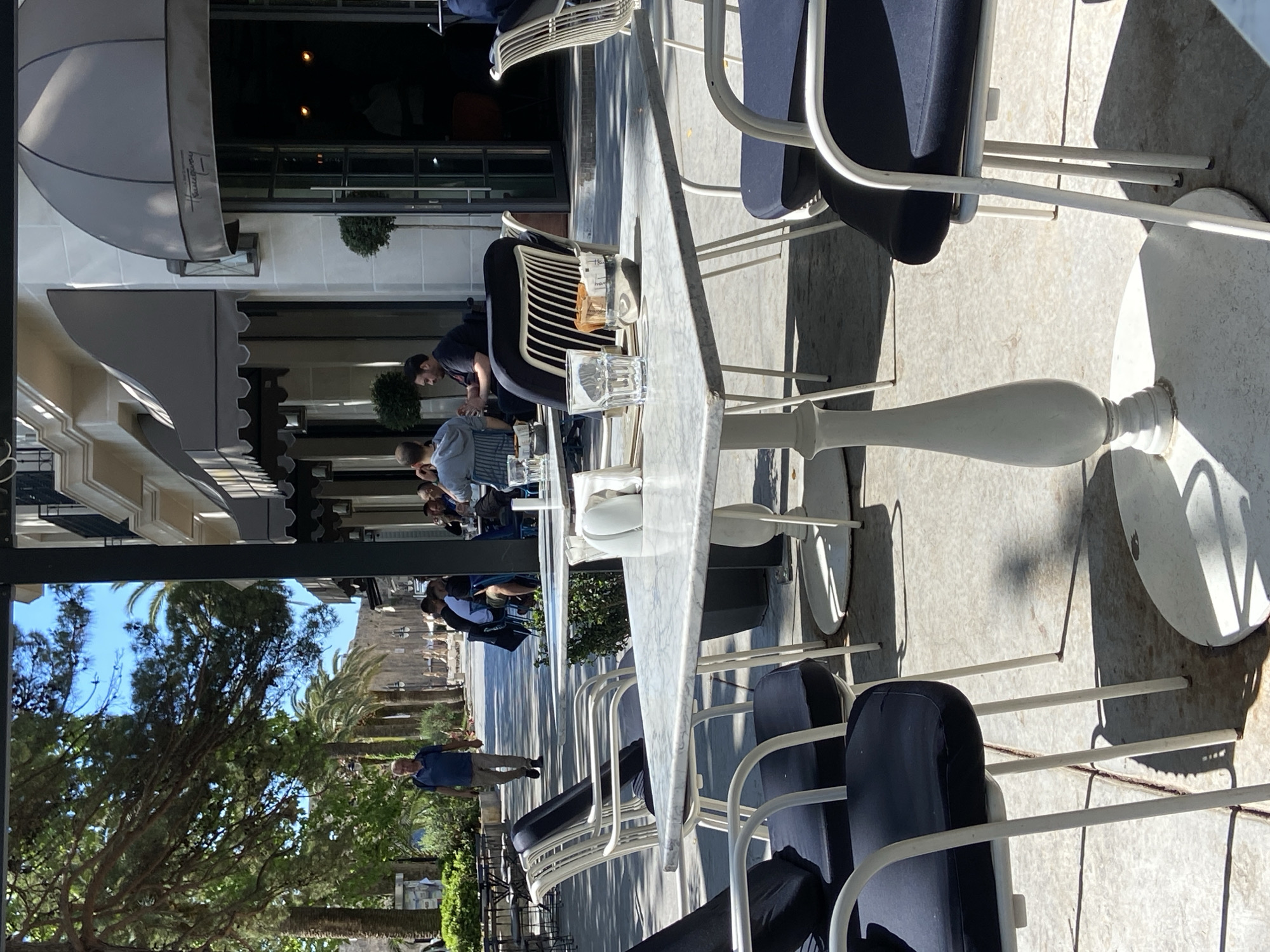 The image depicts an outdoor seating area in Budva, Montenegro. The location features marble-topped tables and white metal chairs with blue cushions, typical of cafes or restaurants. Patrons are seated at some tables, with one man in a blue shirt walking on the pedestrian way. The scene is sunny, with tree-lined street stretching beyond the immediate seating. Architectural details show awnings and facades characteristic of the city's building style. Glasses and drinks are visible on the table.