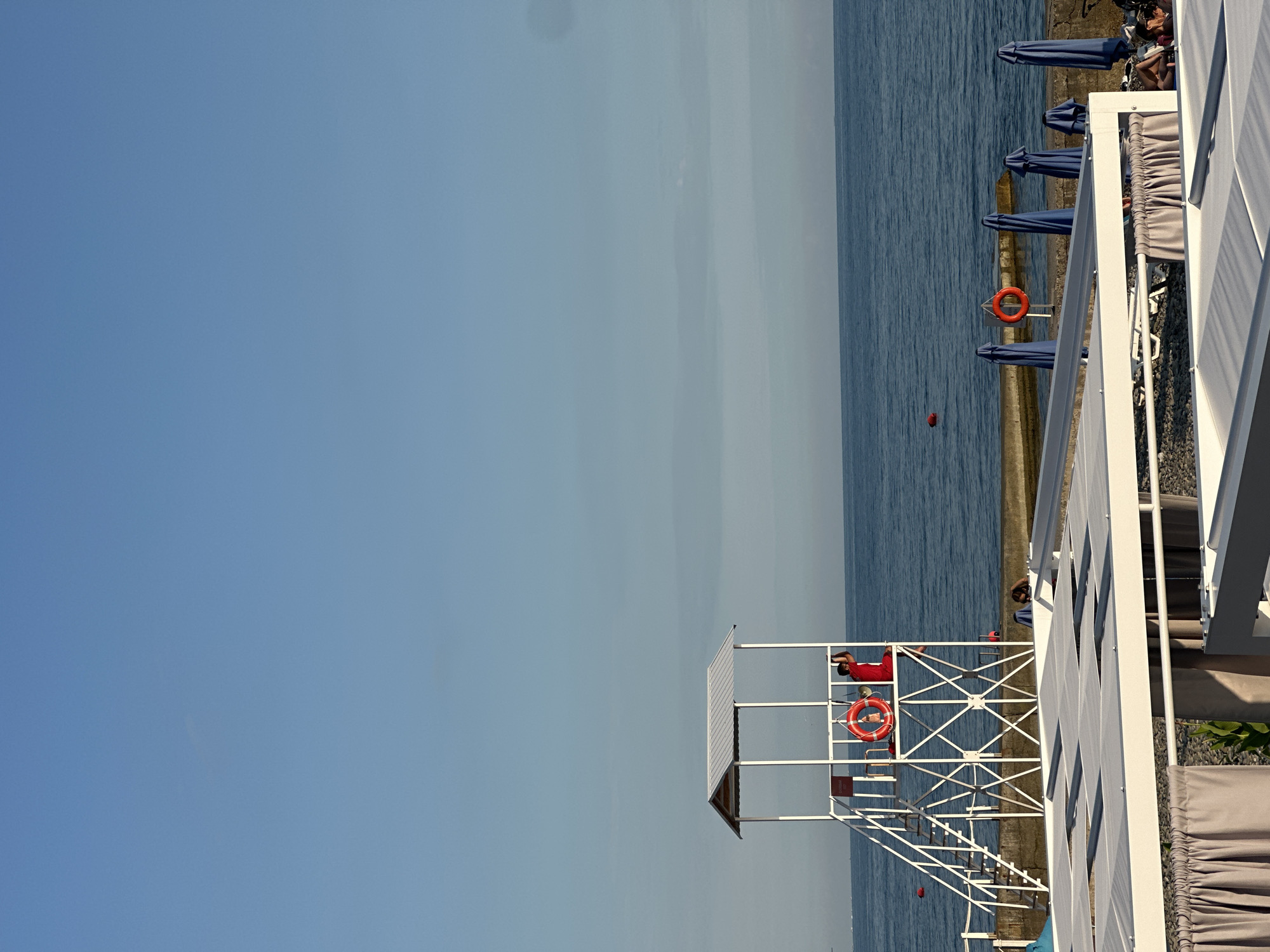 The image depicts a scene on a beach in Khostinskiy, Russia. In the foreground, there are white structures with grey curtains, possibly part of a beach club or seating area. Beyond these, the sea stretches out under a clear blue sky with a few faint clouds. A concrete walkway runs along the edge of the sea.
Dominating the mid-ground is a white lifeguard tower with a red life preserver hanging on its railing. A person in a red uniform is standing on the tower, appearing to be a lifeguard. Further along the walkway, another red life preserver is mounted on a post. Several closed blue umbrellas are arranged near the water's edge. A red buoy is visible in the sea.
In the background, the horizon line is clearly defined by the meeting of the sea and the sky. The overall atmosphere is that of a calm day at a seaside resort.