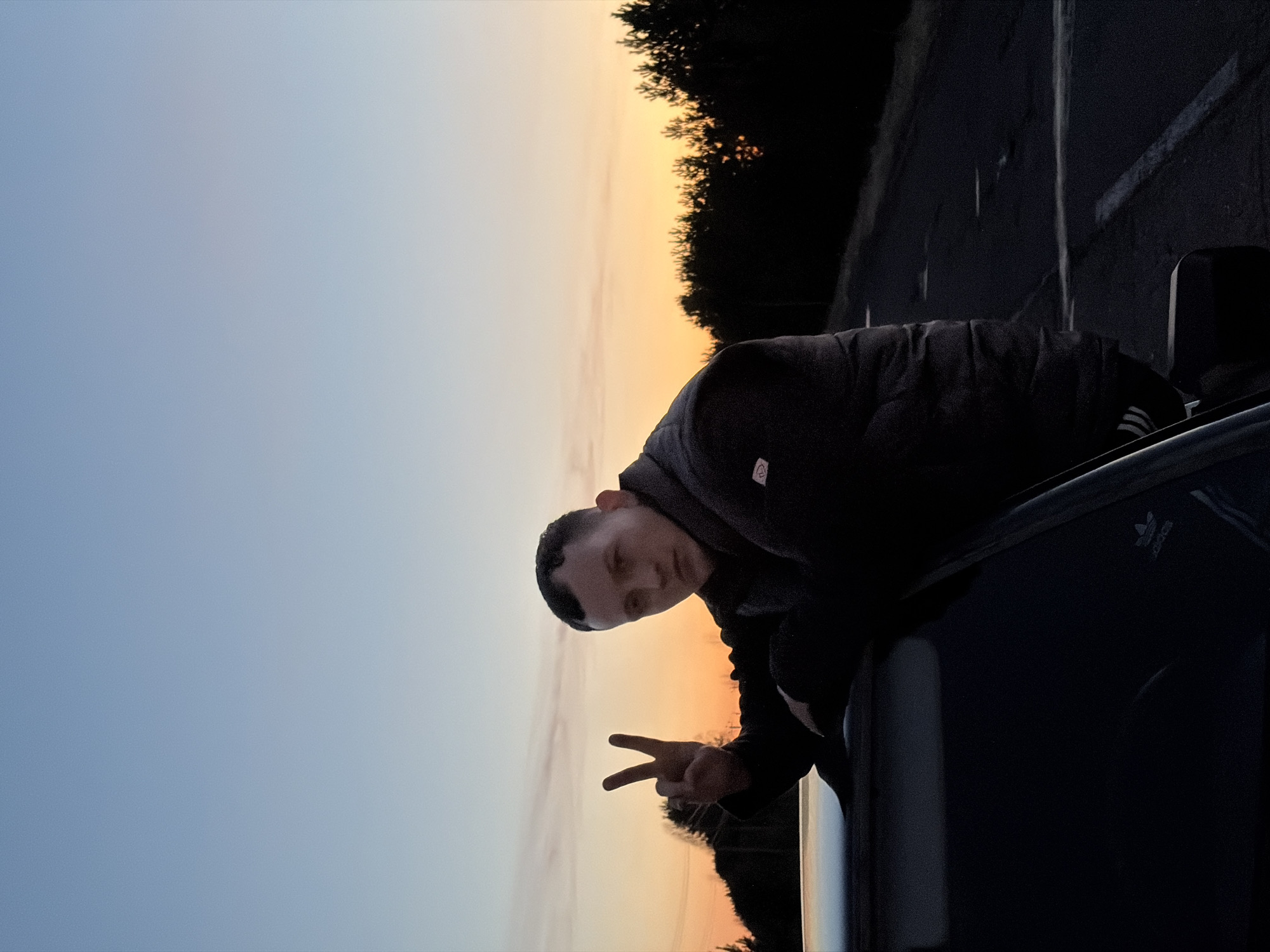 A young person, likely male, is captured leaning out of a car window, making a peace sign with their right hand. The individual is wearing a dark, puffy jacket, a grey hooded sweatshirt underneath, and a white headband. Their gaze is directed towards the camera with a neutral to slightly smiling expression. The setting is outdoors during sunset or dusk, indicated by the sky's gradient, which transitions from a clear, light blue at the top to a warm orange and yellow hue near the horizon. Silhouetted trees line the horizon, suggesting a road or open area, consistent with a location like Kharnavy, Russia. The car, dark in color, is partially visible in the lower right, with an "Adidas" logo discernable on what appears to be the car's interior, possibly a seat or trim. The low light of the evening casts much of the foreground and the person's attire into shadow, creating a backlit effect against the colorful sky.