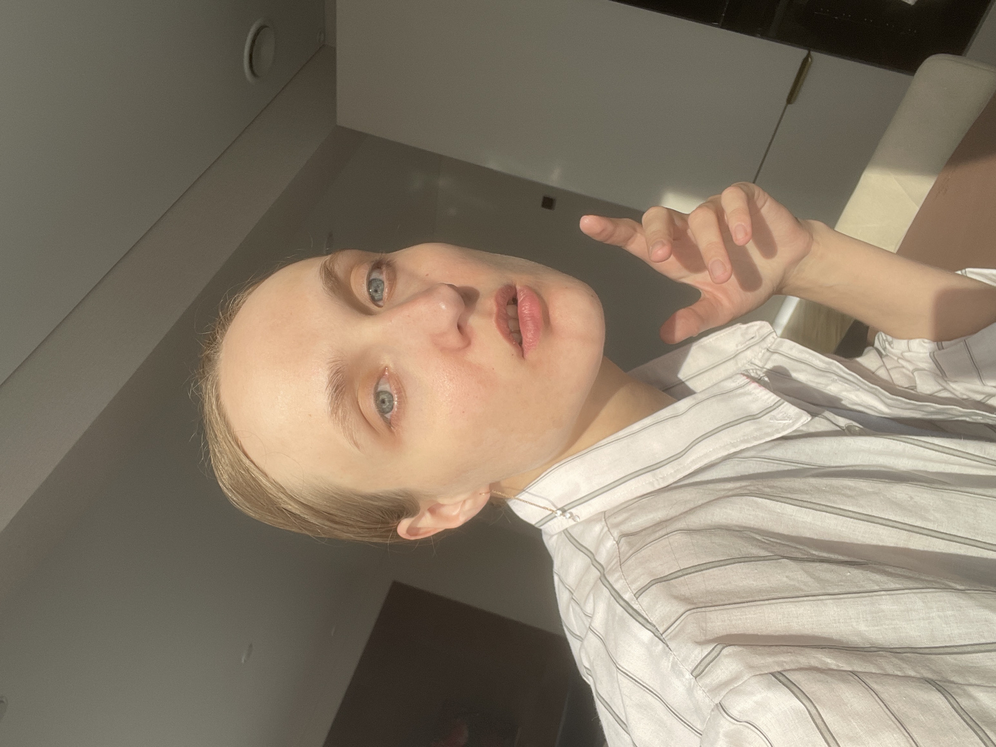 A light-skinned person with light hair is posing indoors, wearing a white and gray striped collared shirt. The person's face is prominently featured, revealing blue eyes and pink lips. The person's right hand is raised, fingers slightly spread. The interior space includes light-colored walls and cabinets. The photograph may have been taken near Sepych, Russia, but there's not enough information in the image to confirm this.