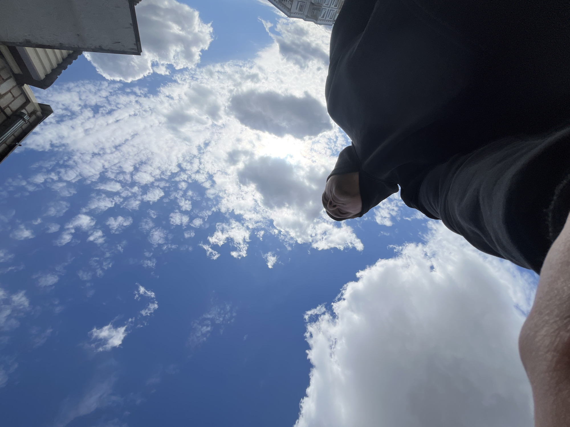 The image is a low-angle shot looking upwards. The sky is mostly blue with scattered white clouds of varying sizes and shapes. The sun is partially obscured by one of the cloud formations. A portion of a building is visible at the upper left edge, suggesting an urban environment. A person wearing a black hooded garment is positioned on the right side of the frame. The person's face and part of their arm are visible. There is no way to confirm that the image was taken in Vysokovsk, Russia from the provided information.