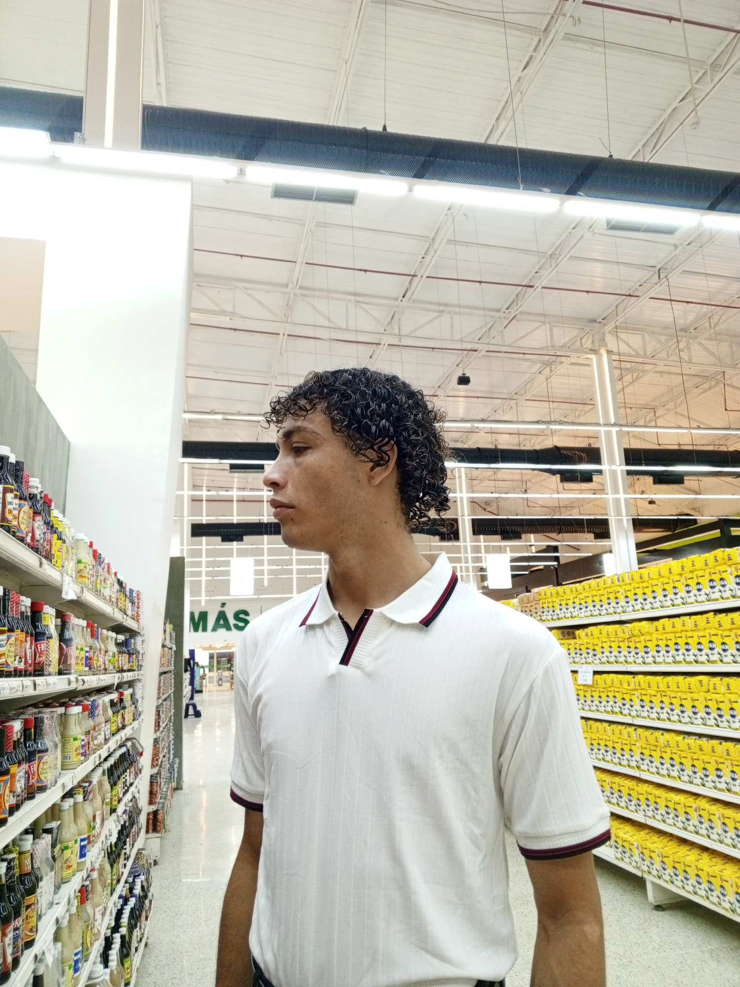 A medium shot depicts a young man with curly, dark brown hair standing in an aisle of a grocery store. He is wearing a white polo shirt with dark trim on the collar and sleeves. The man is facing left, his gaze directed toward the shelves to his left, which are filled with various sauces and dressings. To his right, shelves are stocked with yellow packages of what appear to be food products. The store has a high ceiling with exposed metal beams and fluorescent lighting. A sign that reads "MAS" is visible in the background. Based on the sign, it is likely the location is an aisle of the supermarket MAS x MENOS in the city of Porlamar, Venezuela.