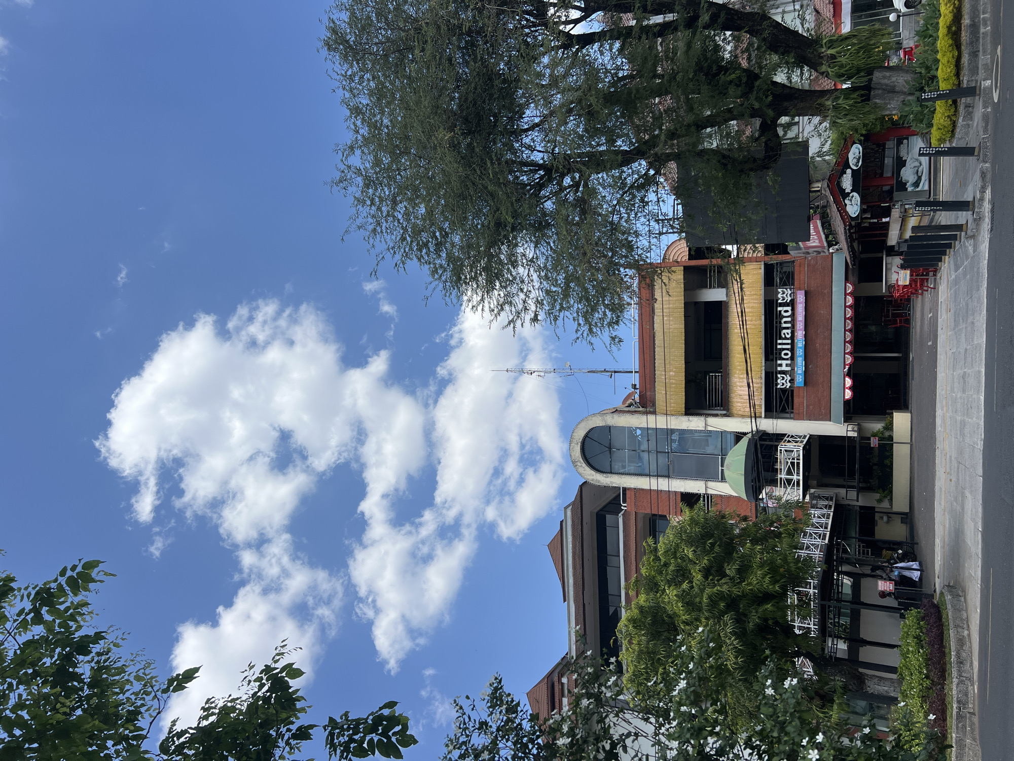 The image captures a street view in Yogyakarta, Indonesia, under a bright blue sky dotted with fluffy white clouds. In the foreground, a paved road runs parallel to a sidewalk lined with bollards. Beyond the sidewalk, a low-lying garden bed with green and purple foliage separates the sidewalk from a multi-story building.
The building itself is a mix of architectural styles. The lower floors feature a commercial facade with shops and restaurants. One prominent sign reads "Holland" in large, stylized letters, with smaller text beneath it indicating "BELIVING BELIVING BELIVING." Red outdoor seating with umbrellas is visible in front of this establishment. Further down, another smaller structure with a red roof and decorative elements appears to be a food stall or cafe.
The upper floors of the building are more modern, with large glass windows and balconies. A distinctive arched glass structure protrudes from one section of the building, acting as an elevator or decorative feature. Trees, including a large, leafy tree on the right side of the image, partially obscure the building. In the background, other buildings and a telecommunications tower are visible. The scene appears to be a commercial district in Yogyakarta.